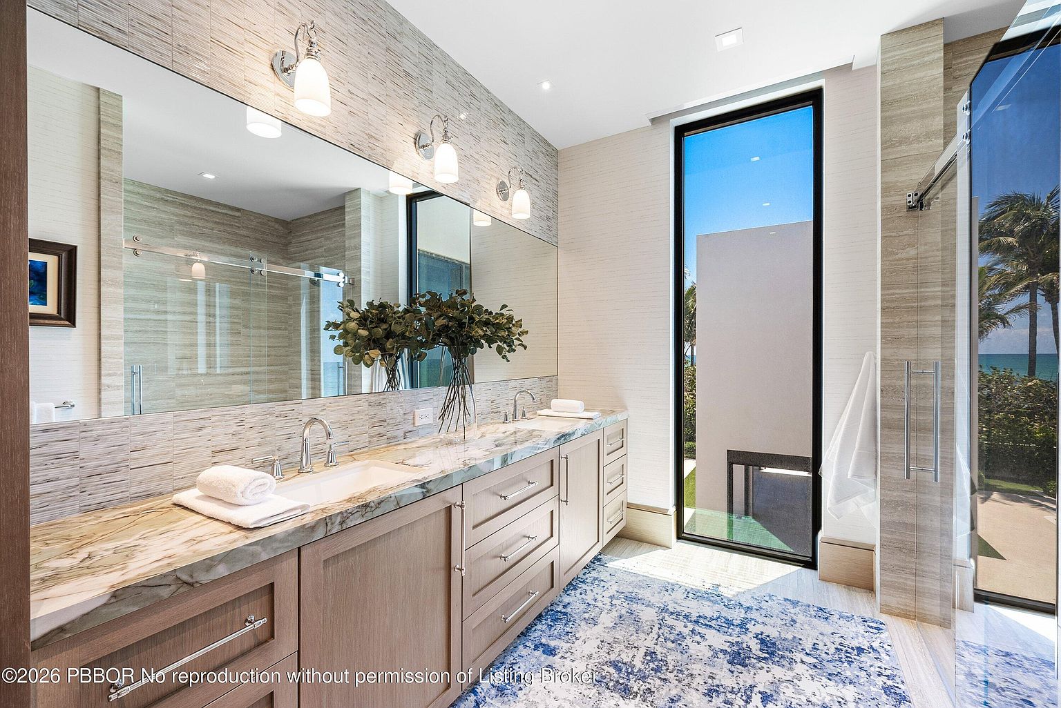 This elegant primary bathroom features a long vanity with a stone countertop, dual sinks, and light wood cabinetry. A large mirror spans the wall above the vanity, reflecting the space and the tall window that provides a view of the outdoors. The room is accented by a blue and white patterned rug and a glass-enclosed shower, creating a bright and sophisticated atmosphere.