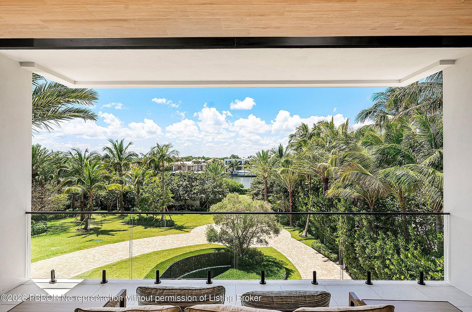 This elevated balcony offers a panoramic view of a lush, tropical landscape and a serene waterway in the distance. The space features a sleek, frameless glass railing that ensures an unobstructed perspective of the manicured lawn, curved stone pathways, and mature palm trees below. The overhead structure is finished with a light wood-toned ceiling, creating a sophisticated and airy outdoor living area.