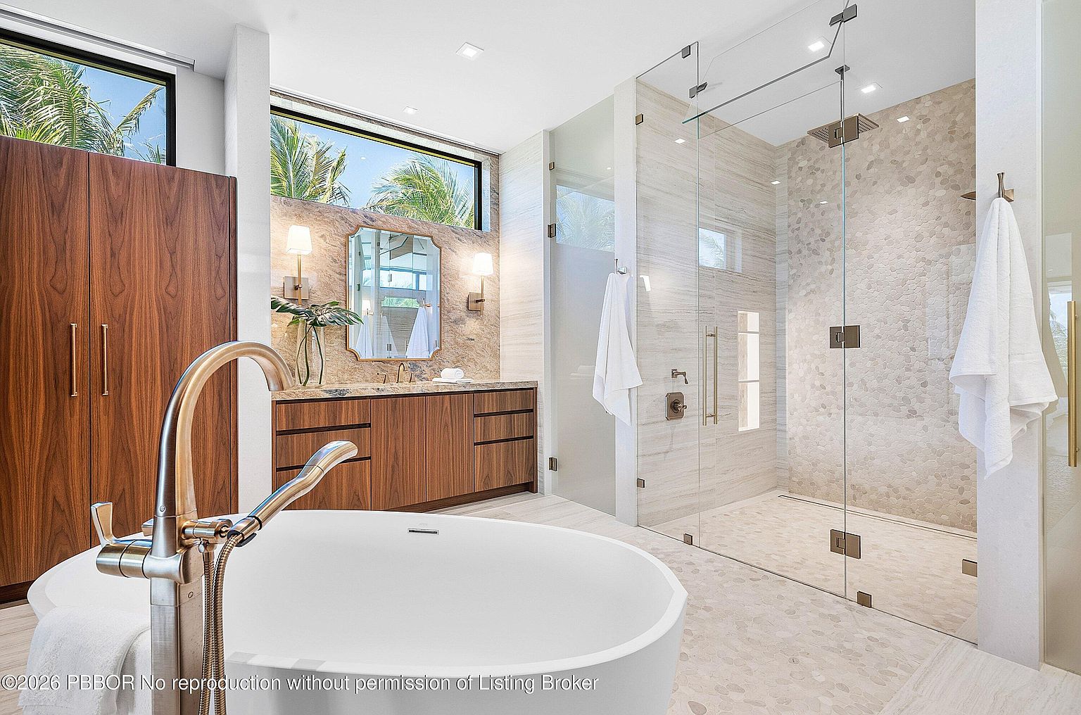 This luxurious primary bathroom features a freestanding soaking tub in the foreground and a spacious walk-in shower with a glass enclosure and pebble-textured flooring. A floating wood vanity with a stone countertop sits beneath a decorative mirror and tall windows that allow for natural light. The space is finished with neutral-toned wall tiles and modern fixtures, creating a serene and high-end spa-like atmosphere.