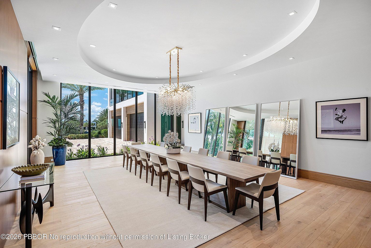This expansive dining room features a long, light wood table surrounded by modern, neutral-toned chairs, positioned beneath a striking, multi-tiered crystal chandelier. The room is defined by a large circular recessed ceiling detail and floor-to-ceiling windows that provide an unobstructed view of the outdoor patio and landscaping. Three large, leaning mirrors reflect the space, enhancing the sense of scale and light within this sophisticated, contemporary interior.