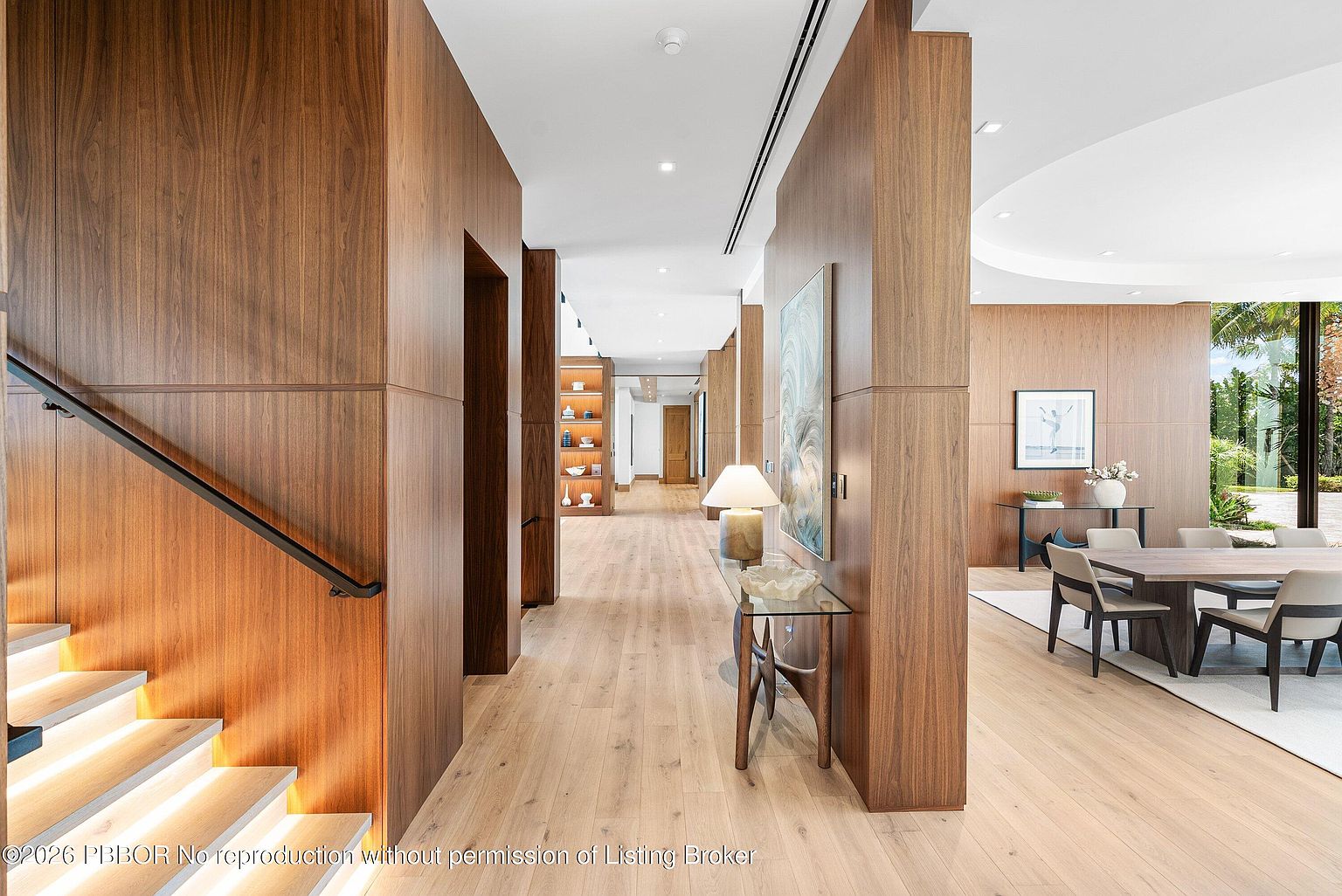 This elegant interior view captures a spacious hallway featuring warm wood-paneled walls and light-toned wood flooring. To the left, a staircase with a modern railing leads upward, while the hallway extends toward a dining area on the right, furnished with a contemporary table and chairs. The perspective emphasizes the open, airy flow of the home, highlighted by recessed lighting and a clean, minimalist aesthetic.