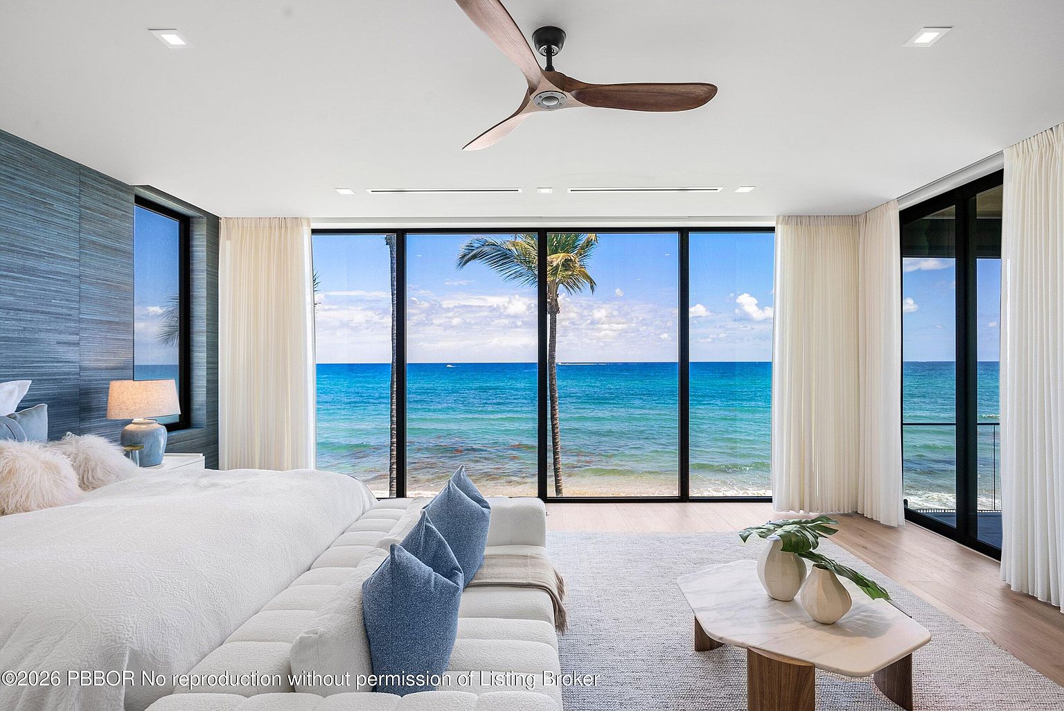 This luxurious primary bedroom features a minimalist aesthetic with a large, plush white bed positioned to face expansive floor-to-ceiling windows that offer a panoramic view of the ocean. The room is accented by a textured blue feature wall, a modern wooden ceiling fan, and a light-toned area rug over wood flooring. A sculptural coffee table with a plant arrangement sits in the foreground, creating a serene and sophisticated atmosphere.