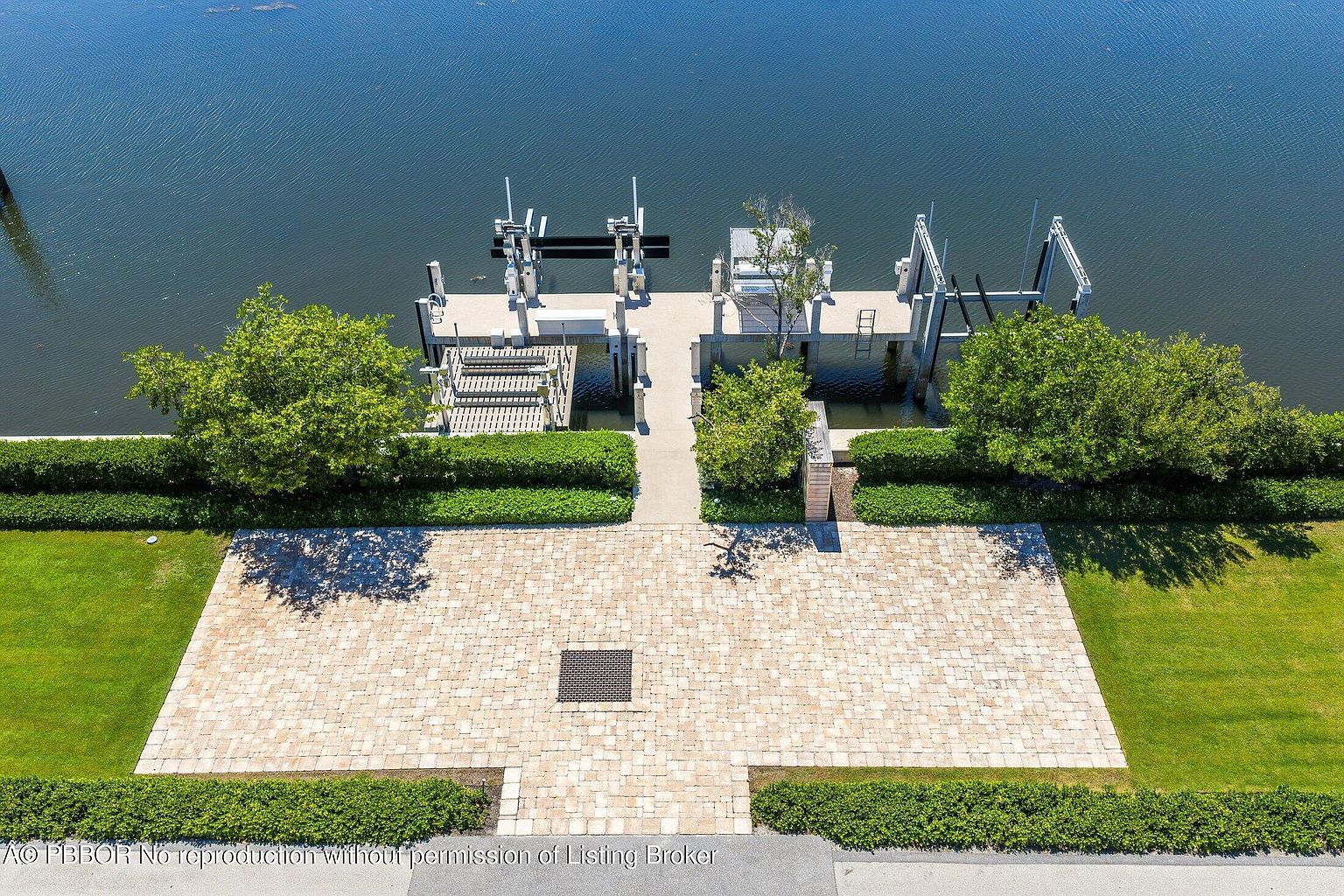 This high-angle aerial view captures a beautifully landscaped waterfront property featuring a paved stone patio area that transitions into a manicured lawn. A concrete walkway leads from the patio through a lush green hedge to a private boat dock equipped with multiple boat lifts extending into the calm water. The scene is framed by mature trees and neatly trimmed shrubbery, creating a serene and well-maintained outdoor living space.