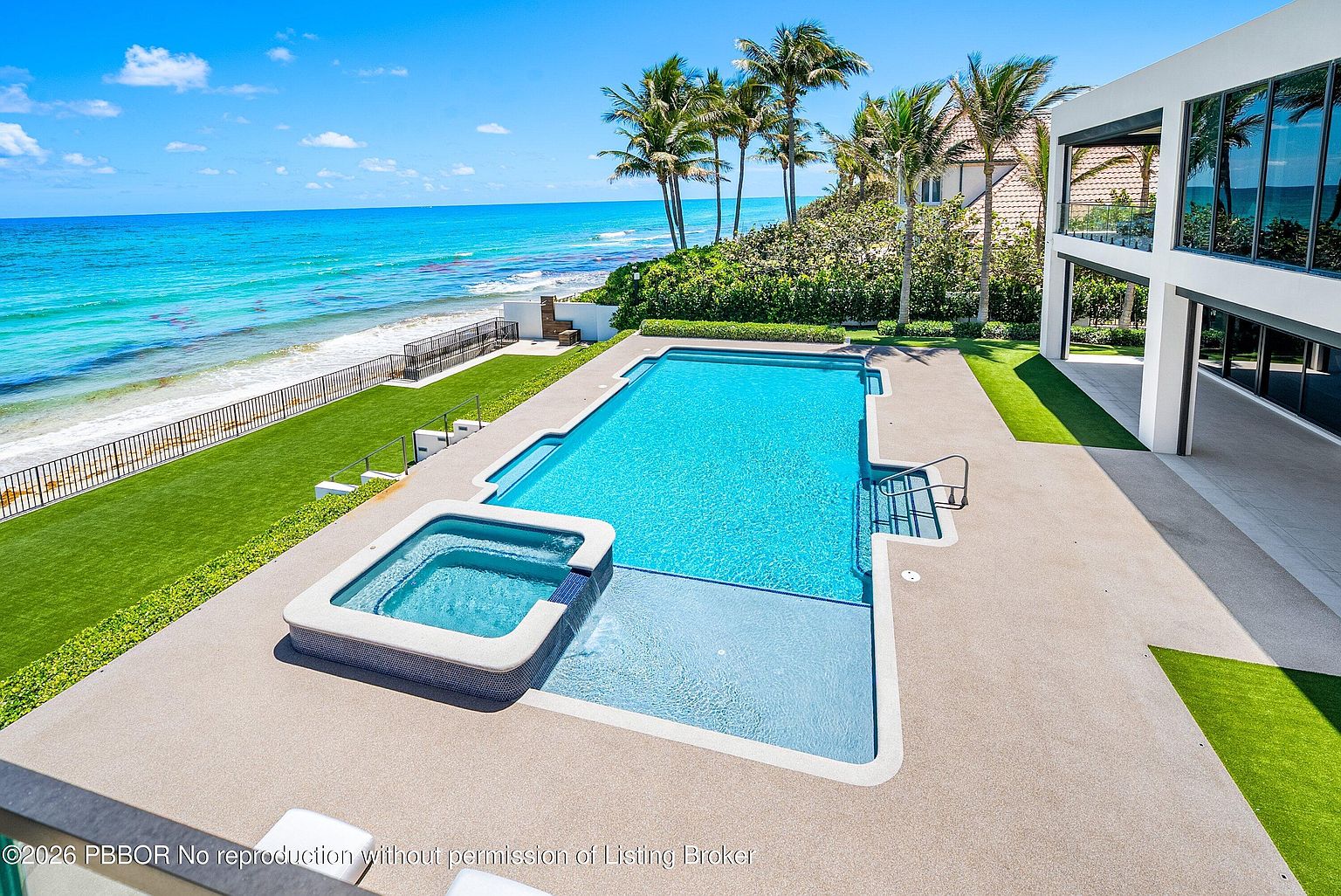 This high-angle aerial view captures a luxurious oceanfront property featuring a modern rectangular swimming pool with an integrated spa. The pool deck is finished in a textured light-colored material, complemented by a manicured green lawn that stretches toward the turquoise sea. The adjacent contemporary home showcases expansive glass walls, creating a seamless transition between the indoor living spaces and the outdoor coastal environment.