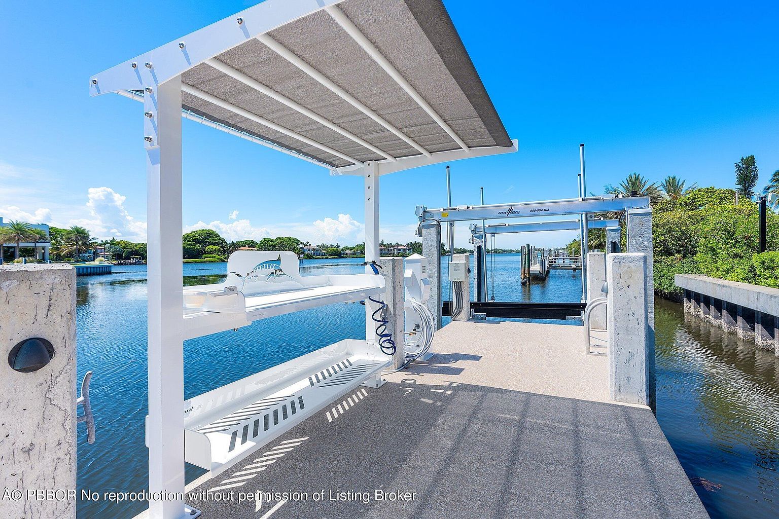 This image showcases a private boat dock extending over a calm, blue waterway, featuring a covered seating area with a built-in bench adorned with a marlin graphic. The dock is equipped with a professional boat lift system and a coiled hose, highlighting its functionality for watercraft enthusiasts. The perspective is from the dock looking out toward the water, capturing a bright, sunny day with lush green vegetation lining the distant shore.