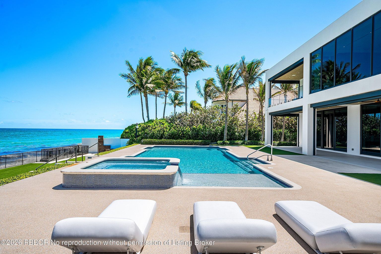 This luxurious oceanfront outdoor space features a pristine swimming pool and integrated spa, set against a backdrop of the turquoise sea. The modern white architecture of the home provides a clean contrast to the lush palm trees and manicured landscaping. In the foreground, three white lounge chairs are positioned on a textured deck, creating an inviting atmosphere for relaxation and coastal living.