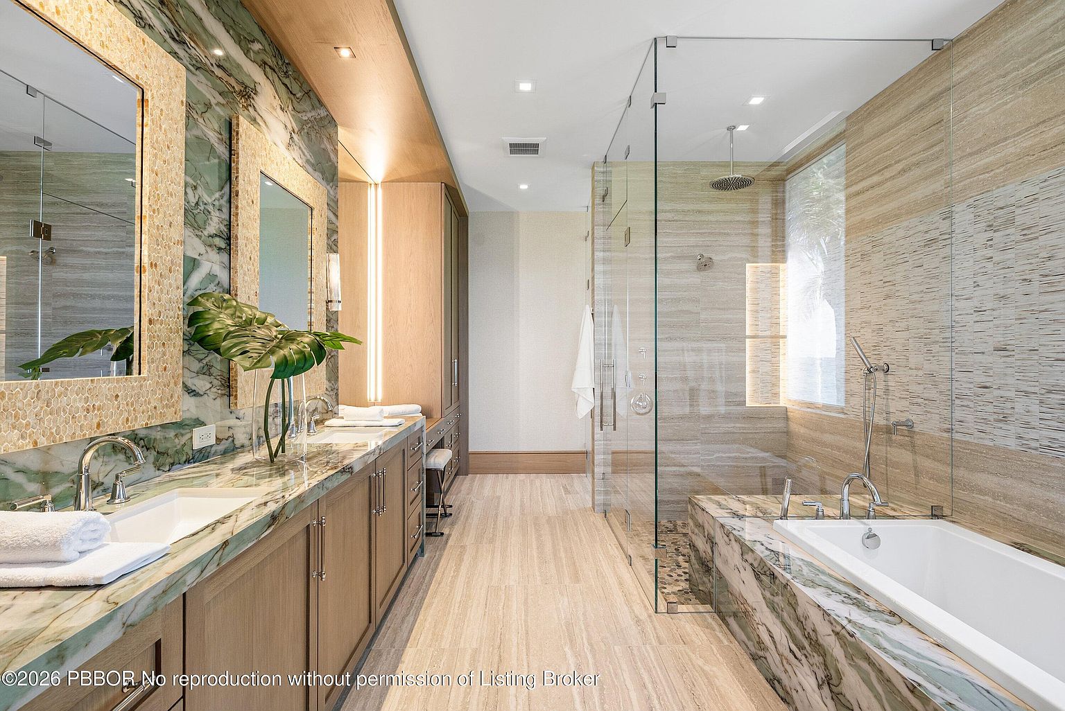 This luxurious primary bathroom features a long vanity with dual sinks set into a patterned stone countertop, complemented by matching stone wall accents and dual mirrors. To the right, a glass-enclosed wet room houses both a rainfall shower and a deep soaking tub, all finished with neutral-toned wall tiles. The space is illuminated by recessed lighting and warm accent lighting, creating a sophisticated and spa-like atmosphere.