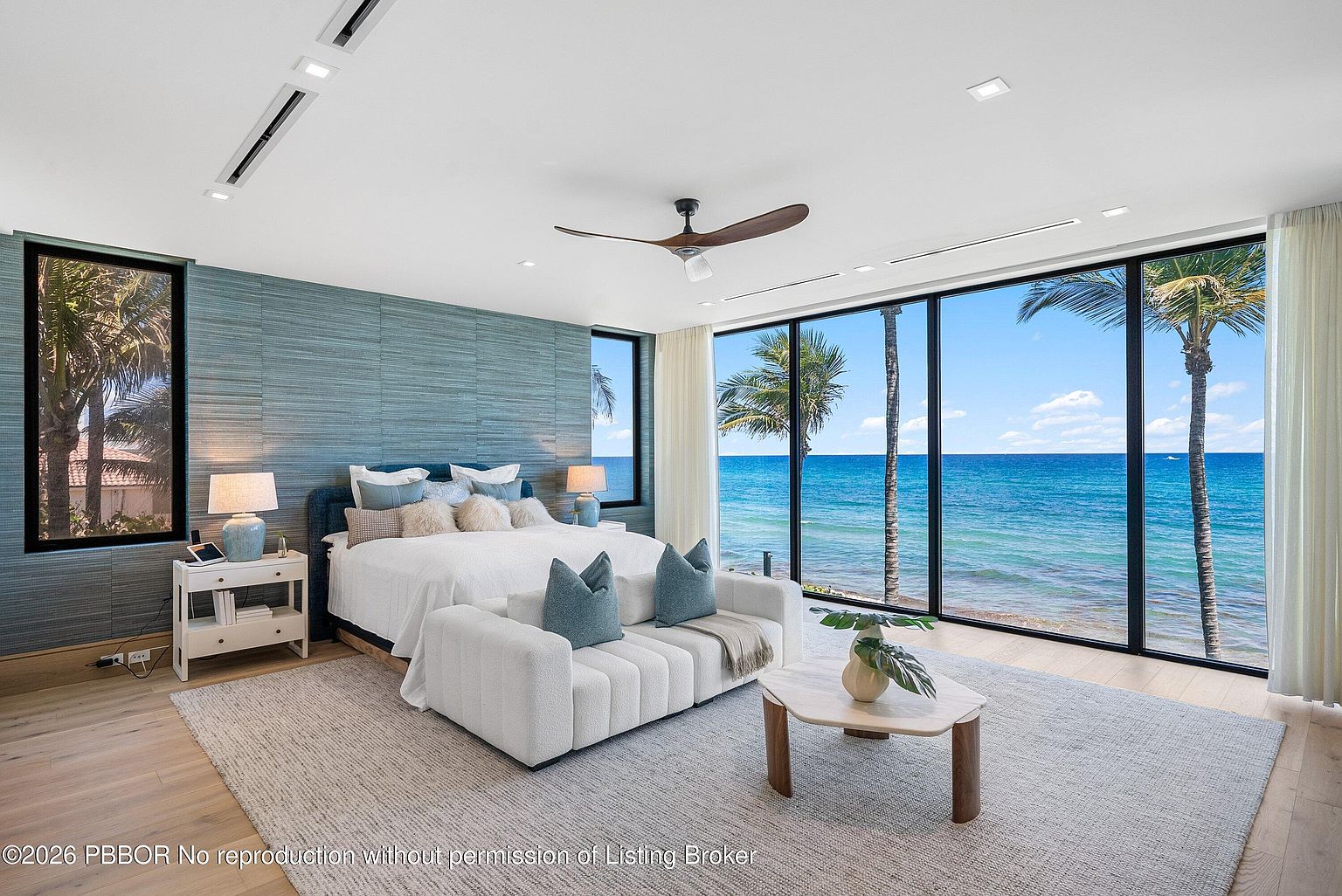 This luxurious primary bedroom features a textured blue accent wall and a plush bed centered in the room, facing expansive floor-to-ceiling windows that offer panoramic ocean views. A modern white sofa sits at the foot of the bed, complemented by a light wood coffee table and a neutral area rug. The room is bathed in natural light, creating a serene and sophisticated atmosphere that emphasizes the connection to the coastal landscape.