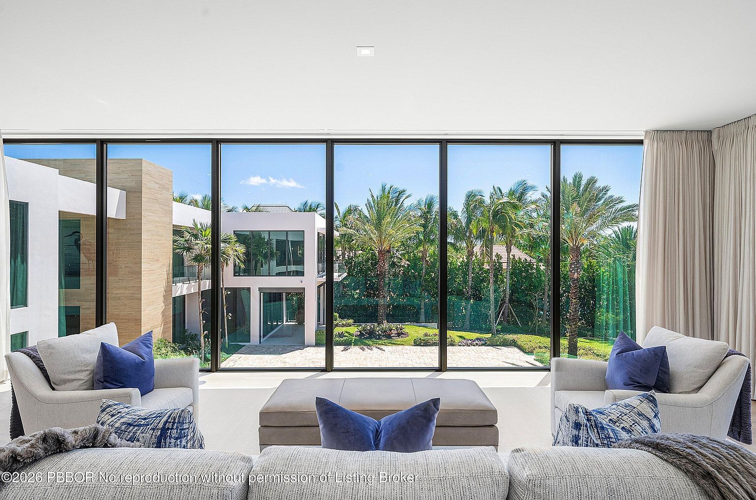 This bright and airy living room features a neutral color palette with plush seating arranged around a central ottoman. The space is defined by expansive floor-to-ceiling windows that provide a seamless transition to the outdoor landscape, showcasing a manicured garden and a modern neighboring structure. The minimalist design emphasizes the natural light and the serene, verdant view beyond the glass.