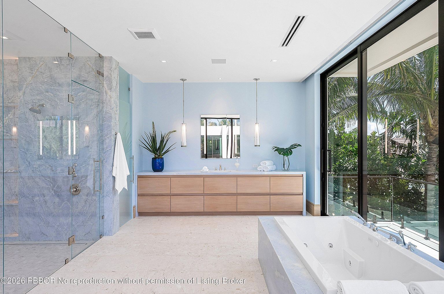 This spacious primary bathroom features a light blue color palette, a long vanity with light wood cabinetry, and a large walk-in shower enclosed by glass panels with blue-toned stone walls. A deep soaking tub sits near floor-to-ceiling windows that provide a view of lush tropical greenery outside. The room is illuminated by sleek pendant lights and recessed ceiling lighting, creating a serene and luxurious atmosphere.