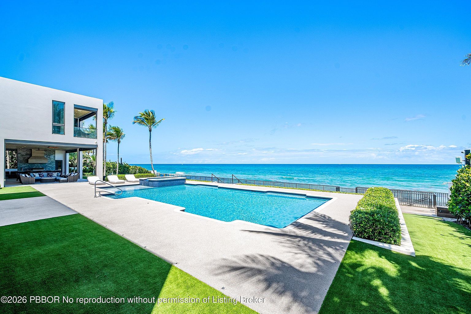 This luxurious oceanfront property features a sprawling rectangular swimming pool with an integrated spa, set against a backdrop of the vast blue sea. A modern, minimalist home with a covered outdoor lounge area sits to the left, while a manicured green lawn and a neat hedge border the pool deck. The scene is captured from a slightly elevated perspective, emphasizing the seamless transition between the private outdoor living space and the expansive coastal horizon.