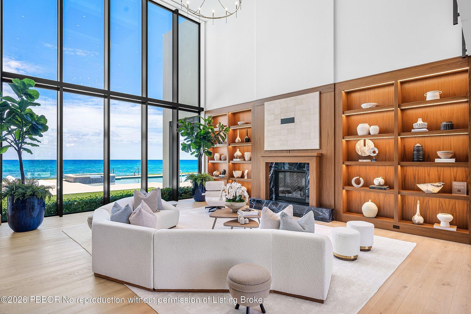 This expansive living room features a curved, light-colored sofa positioned to take advantage of the dramatic, floor-to-ceiling windows that frame a panoramic ocean view. A central fireplace with a dark stone surround serves as a focal point, flanked by custom light wood shelving units filled with curated decor. The room is bathed in natural light, creating an airy and sophisticated atmosphere that seamlessly blends indoor luxury with the coastal landscape.