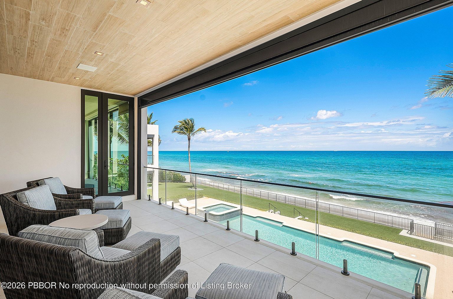 This expansive, covered balcony offers a seamless transition to an outdoor living space with a direct, unobstructed view of the ocean. The area is furnished with comfortable, dark-toned lounge seating and features a sleek glass railing that overlooks a private swimming pool and spa below. The ceiling is finished with light wood paneling, creating a warm contrast against the bright blue sky and turquoise water visible beyond the balcony.