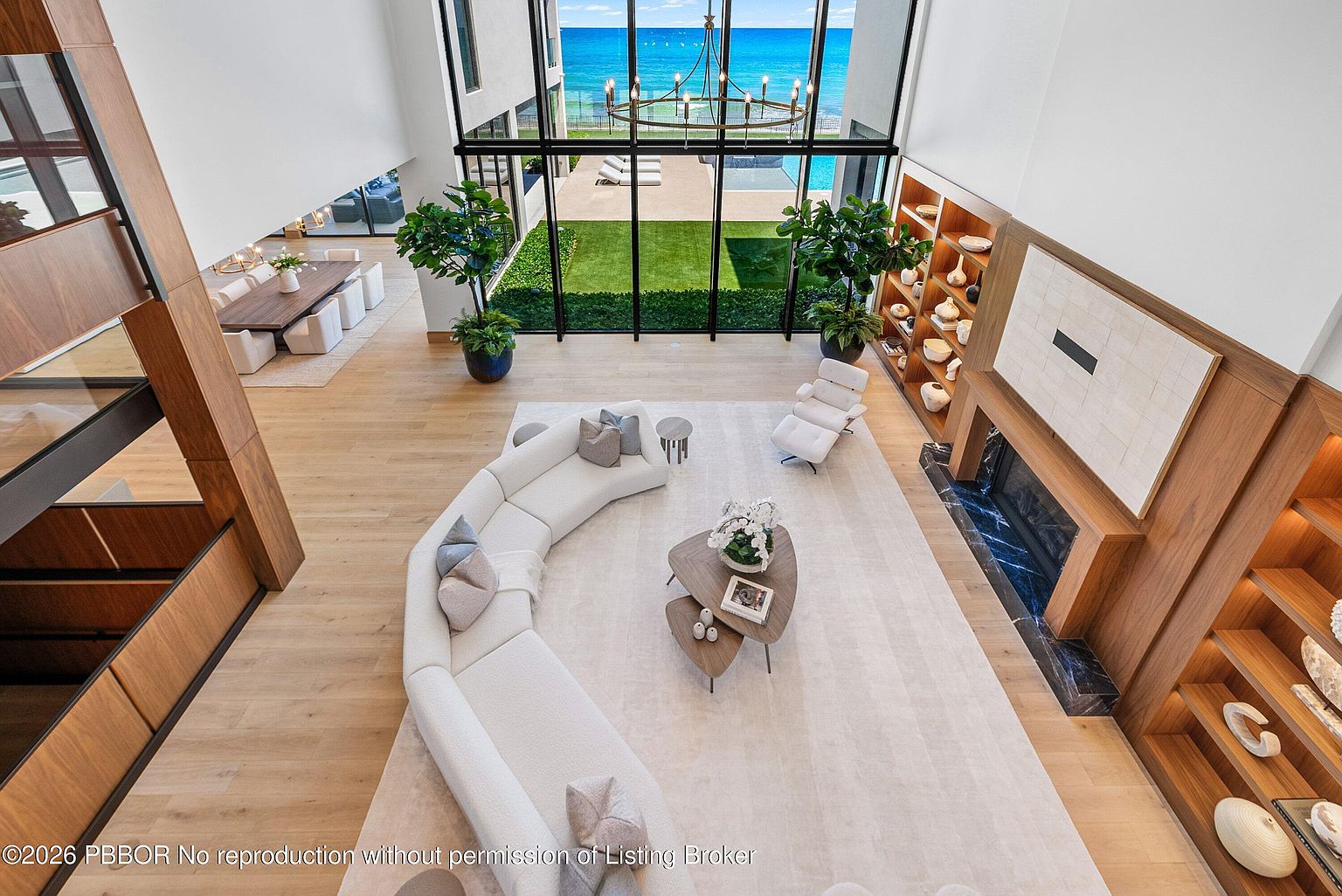 This high-angle shot captures a spacious, open-concept living area characterized by a large, curved white sofa and a minimalist aesthetic. The room features light wood flooring, a tall fireplace with a dark stone surround, and expansive floor-to-ceiling windows that provide a direct view of the ocean and backyard. To the left, a dining area is visible, while built-in shelving units flank the fireplace, creating a sophisticated and airy atmosphere.