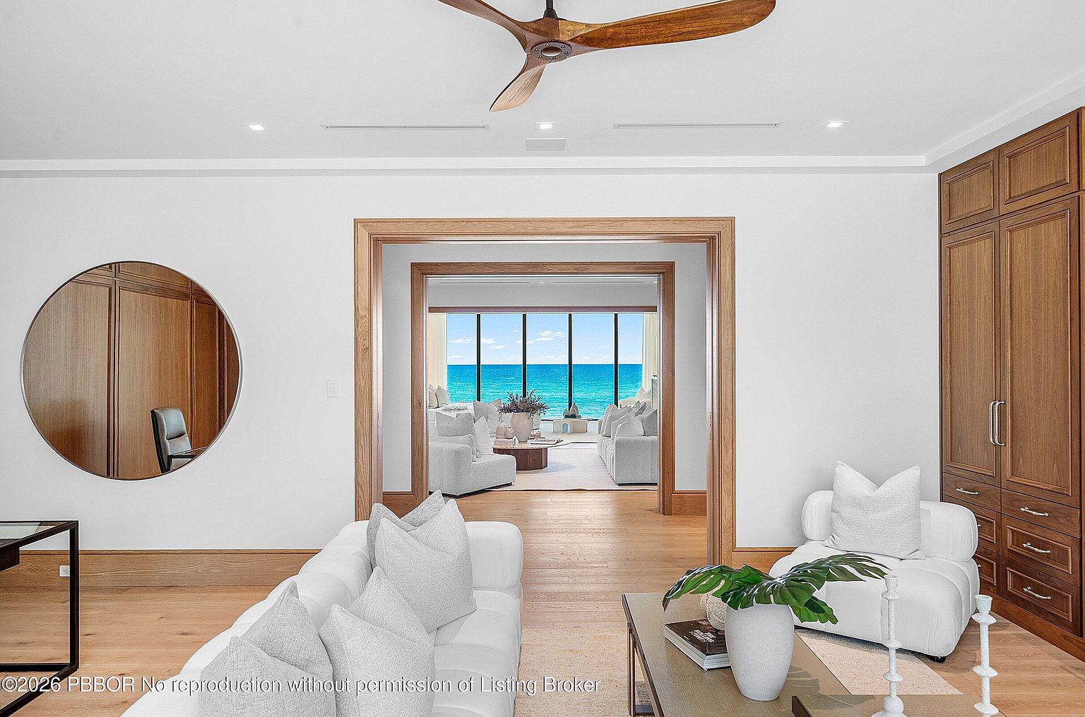 This elegant living area features light wood flooring, a modern ceiling fan, and a large circular wall mirror. The space flows through a wide, wood-trimmed doorway into an adjacent room that showcases expansive windows with a direct view of the ocean. The room is furnished with plush white seating and a coffee table, creating a bright, airy, and sophisticated coastal atmosphere.