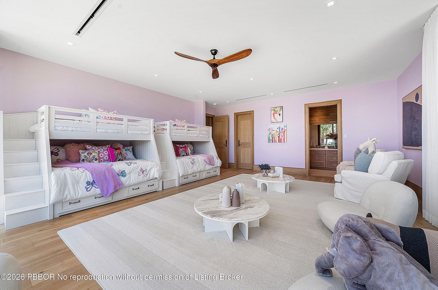 This spacious bedroom features two white bunk beds with integrated staircases, set against soft lavender walls. The room is furnished with two circular stone-topped coffee tables on a large neutral area rug, along with a cozy seating area in the corner. The perspective is a wide-angle shot from the doorway, showcasing the room's generous size and playful, modern aesthetic.