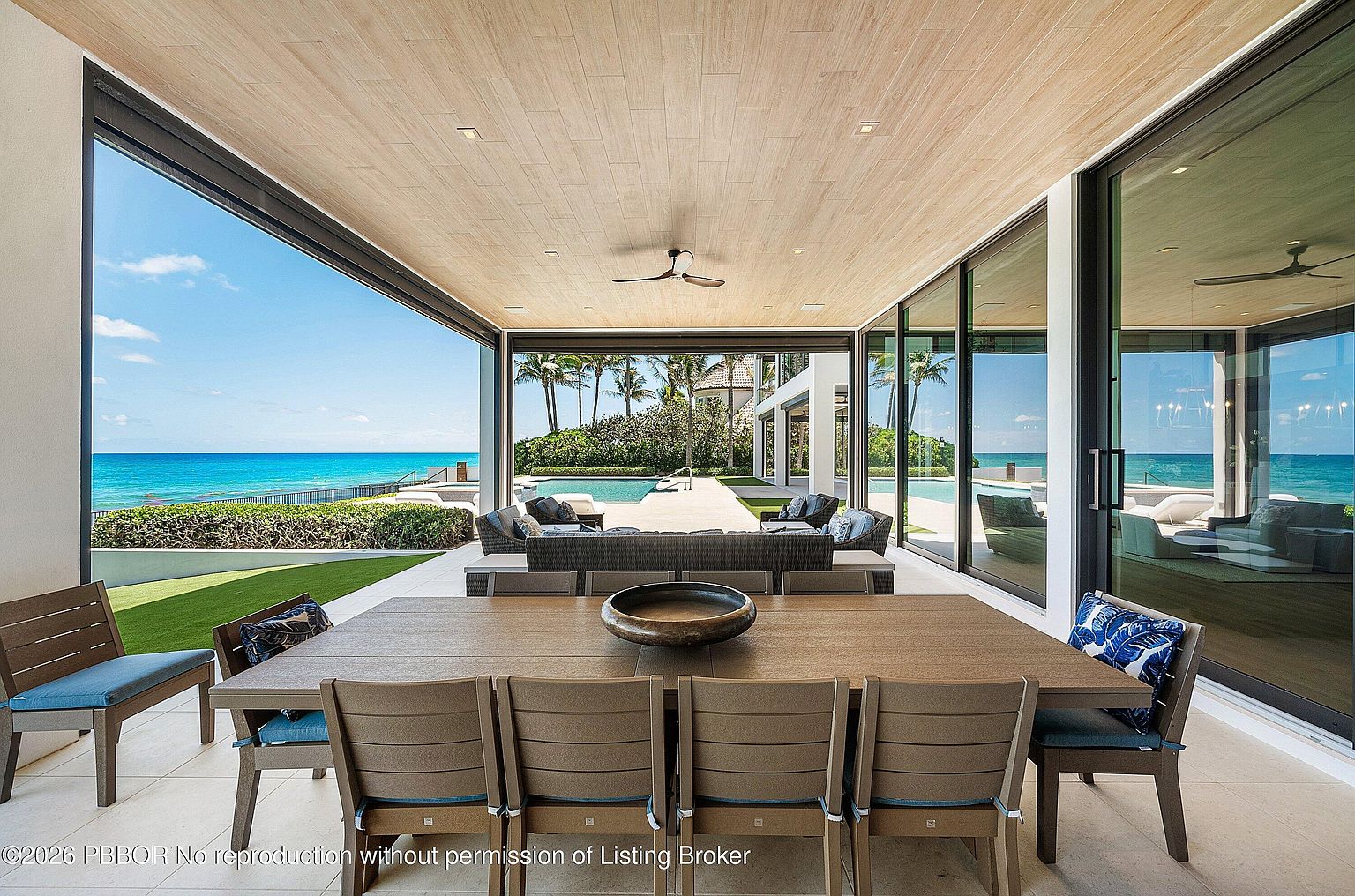 This expansive covered patio offers a seamless transition between indoor and outdoor living, featuring a large dining area with a wooden table and chairs in the foreground. Beyond the dining space, a comfortable lounge area overlooks a pristine swimming pool and the turquoise ocean horizon. The space is defined by a light wood-paneled ceiling and large glass sliding doors that provide an unobstructed, panoramic view of the coastal landscape.
