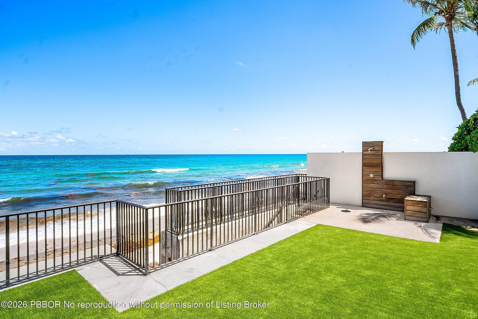 This oceanfront patio area features a manicured artificial turf lawn bordered by a sleek metal railing that overlooks the beach and turquoise sea. A private outdoor shower with a wood-paneled surround and a small wooden bench is integrated into a white privacy wall, offering a luxurious coastal amenity. The perspective captures a wide, bright view of the shoreline, emphasizing the seamless transition between the private outdoor space and the expansive ocean horizon.