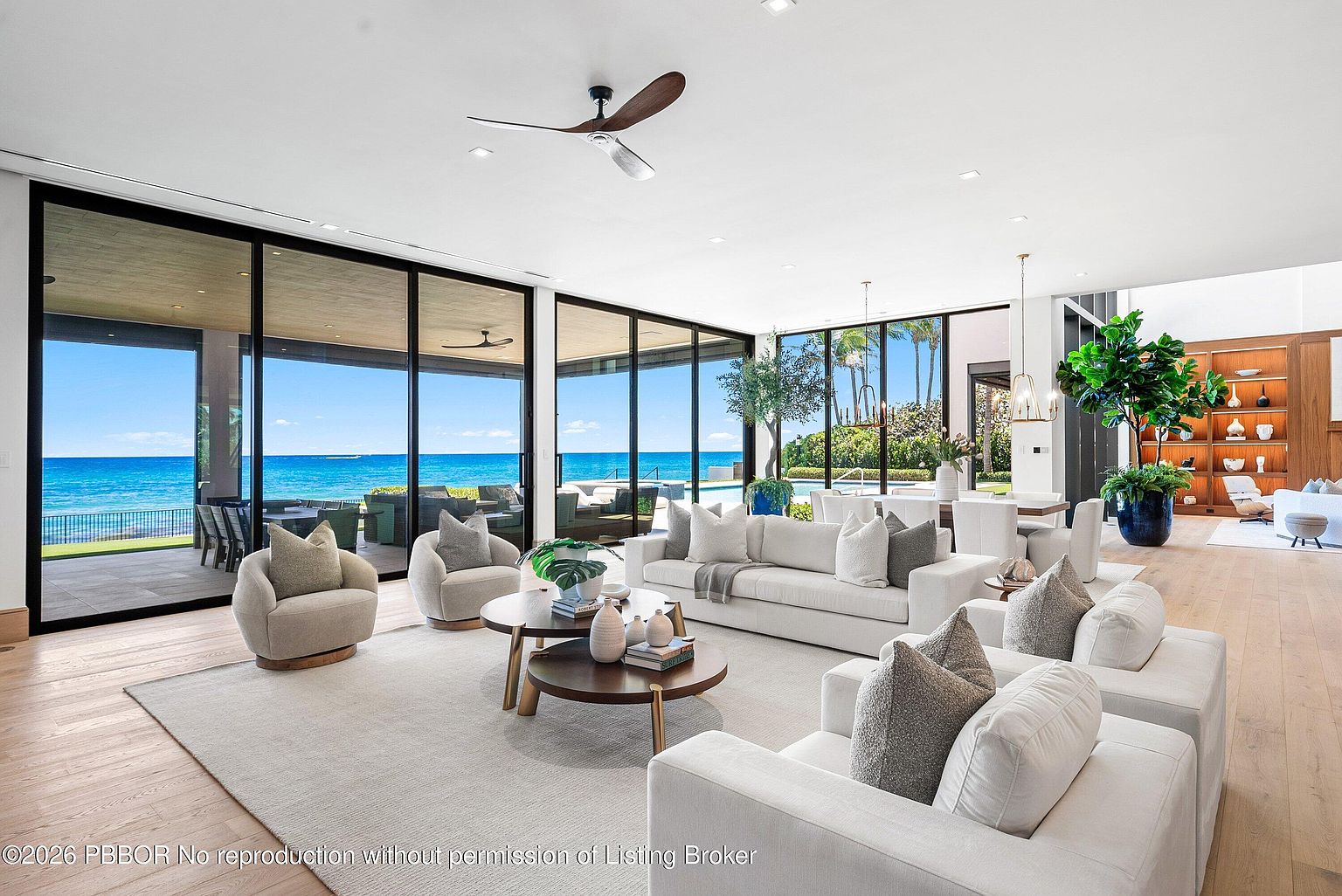 This expansive, modern living room features an open-concept layout with light wood flooring and neutral-toned furnishings. The space is defined by massive floor-to-ceiling glass walls that provide unobstructed, panoramic views of the ocean and a private pool area. A sophisticated seating arrangement with plush sofas and circular coffee tables creates a luxurious atmosphere, complemented by a large potted plant and minimalist decor.