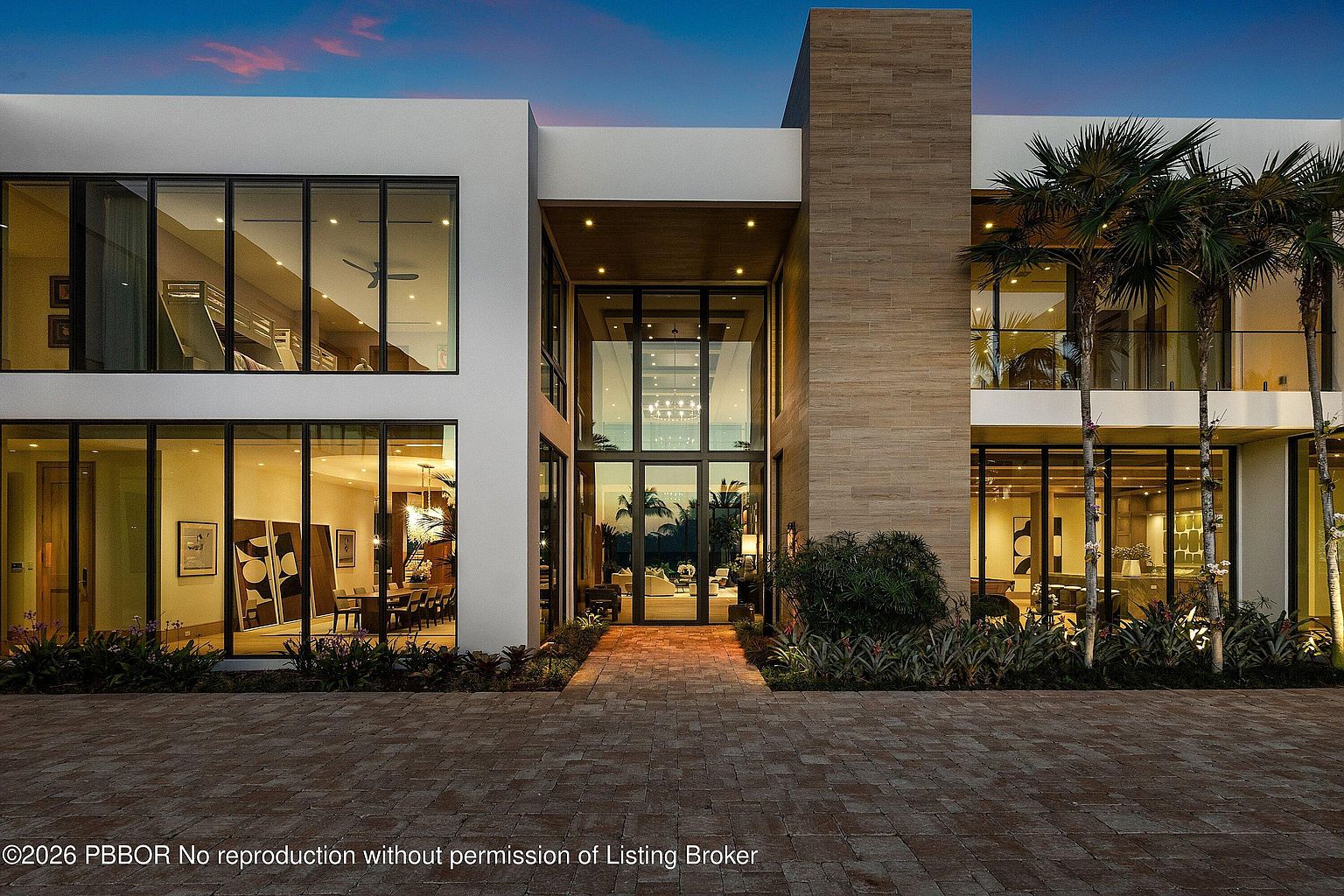 This striking modern residence features a symmetrical design with a prominent central glass entryway and a mix of smooth white walls and textured stone cladding. Large, expansive windows span both levels, creating a seamless connection between the interior living spaces and the exterior environment. The paved driveway leads directly to the grand entrance, framed by manicured landscaping and tall palm trees under a twilight sky.