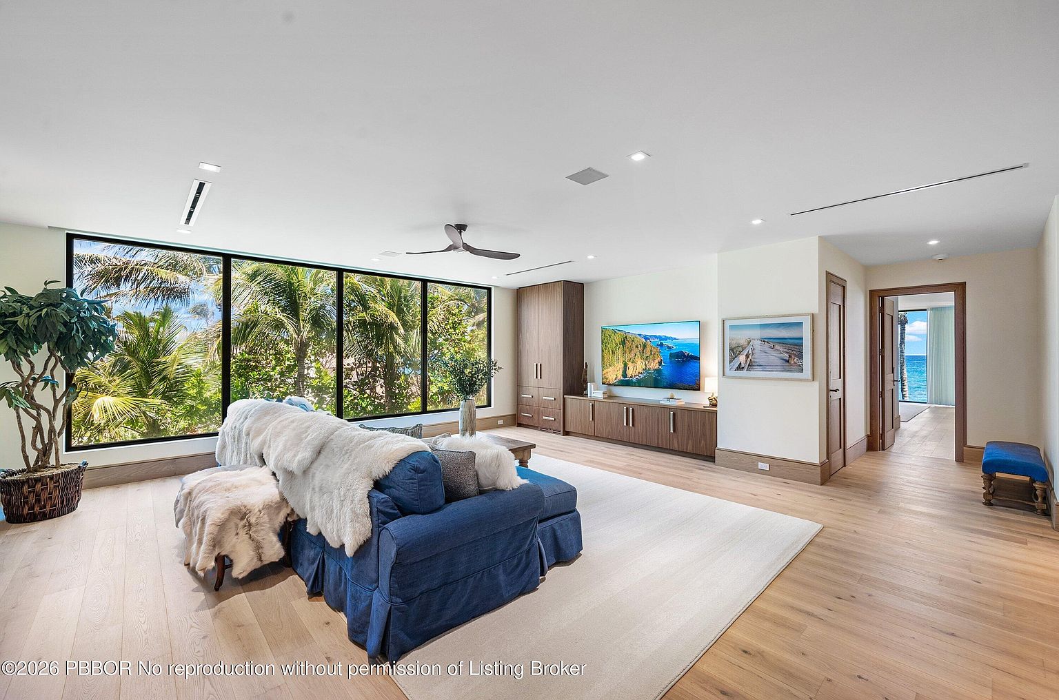 This bright and airy living room features light wood flooring and a large blue sofa adorned with a plush throw blanket. A wall of floor-to-ceiling windows provides expansive views of lush tropical greenery and the ocean beyond, while a media console and tall storage cabinet occupy the opposite wall. The space is finished with a large area rug and a ceiling fan, creating a relaxed, coastal-inspired atmosphere.