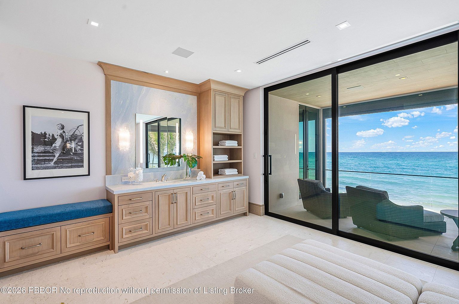 This luxurious primary bathroom features a light wood vanity with a stone countertop and a built-in bench with blue upholstery. The space is defined by floor-to-ceiling glass sliding doors that open onto a private balcony with expansive ocean views. A framed black-and-white photograph adds a sophisticated touch to the serene, light-filled room.