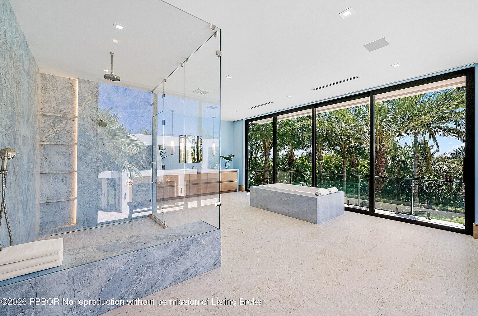 This expansive primary bathroom features a sleek, modern design with a large glass-enclosed shower and a freestanding stone tub positioned near floor-to-ceiling windows. The room is finished with light-toned stone flooring and blue-toned stone wall accents, creating a serene and luxurious atmosphere. The perspective captures the entire space, highlighting the seamless transition between the indoor spa-like amenities and the lush, tropical greenery visible outside.