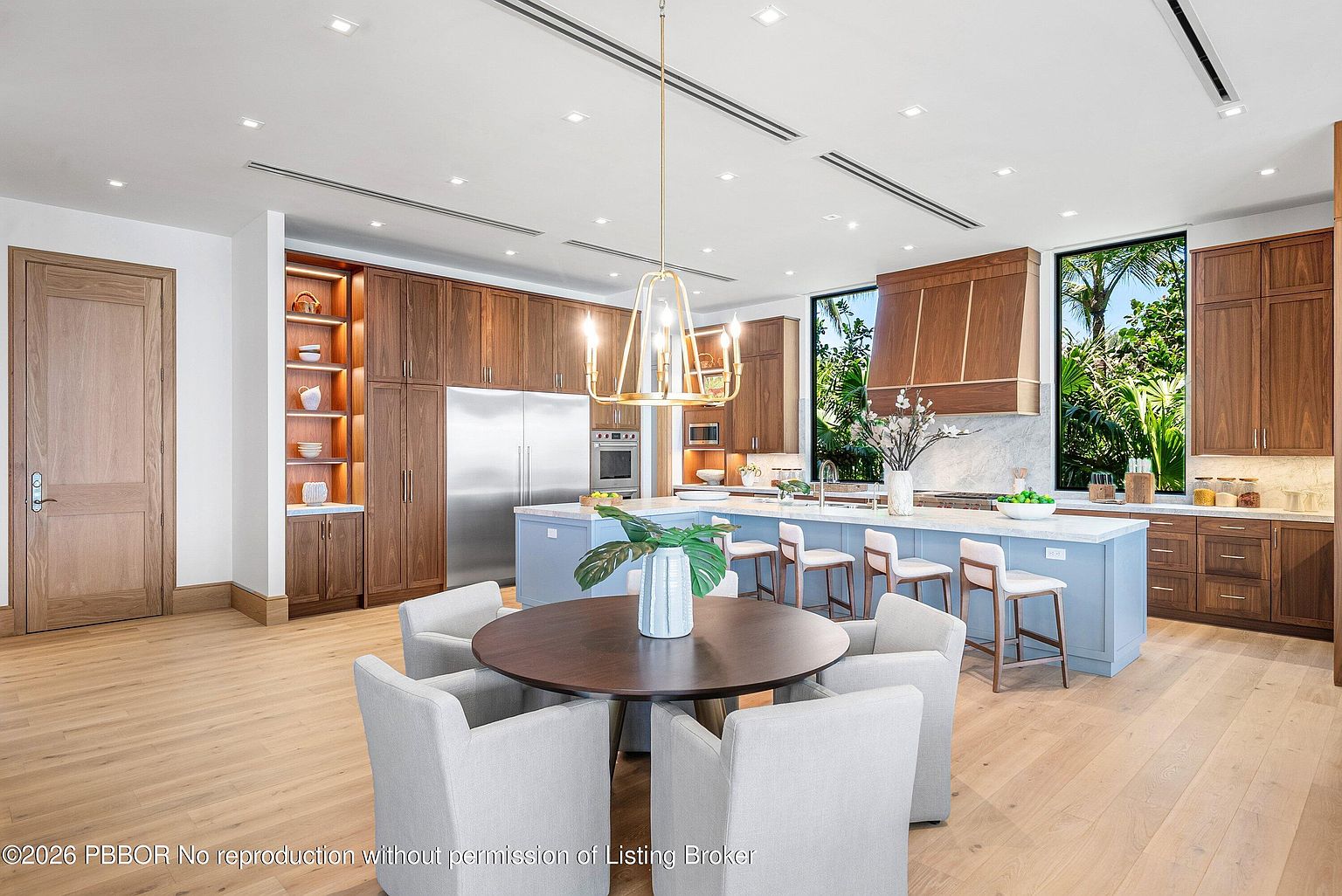 This spacious, open-concept kitchen features a large central island with light blue cabinetry and seating for four, complemented by a round dining table in the foreground. The perimeter is lined with warm-toned wood cabinetry, integrated stainless steel appliances, and open shelving with integrated lighting. Large windows on either side of the range hood provide ample natural light and views of lush greenery, creating a bright and inviting atmosphere.