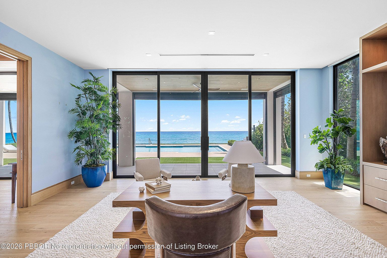 This sophisticated home office features a central wooden desk and a leather chair, positioned to face a set of expansive, floor-to-ceiling sliding glass doors that frame a panoramic ocean view. The room is accented by light blue walls, potted greenery, and a textured area rug over light wood flooring. The seamless transition between the interior workspace and the outdoor pool area creates a serene, high-end atmosphere.