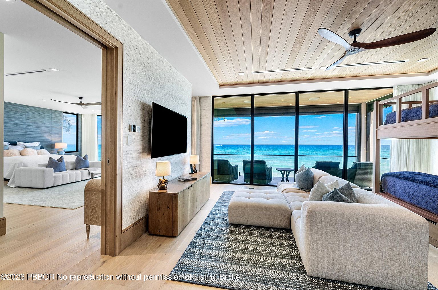 This bright, open-concept living area features a modern aesthetic with light wood flooring and a natural wood-paneled ceiling. A large sectional sofa faces a wall-mounted television, while a bunk bed is tucked into the corner, and a wide doorway leads to an adjacent bedroom. Expansive glass walls provide an unobstructed, panoramic view of the turquoise ocean and a private deck with outdoor seating.