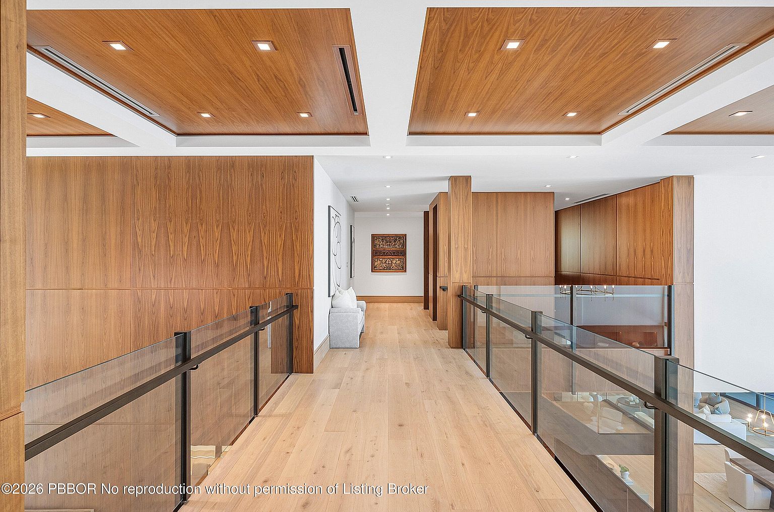 This perspective captures a sophisticated upper-level landing or bridge hallway, characterized by a striking contrast between light wood flooring and warm-toned wood paneling on the walls and recessed ceiling sections. A glass railing provides an open, airy feel, allowing for views into the lower level of the home, while recessed lighting adds a clean, modern aesthetic. The space leads toward a quiet seating area at the end of the hall, creating an elegant and well-lit transition space.