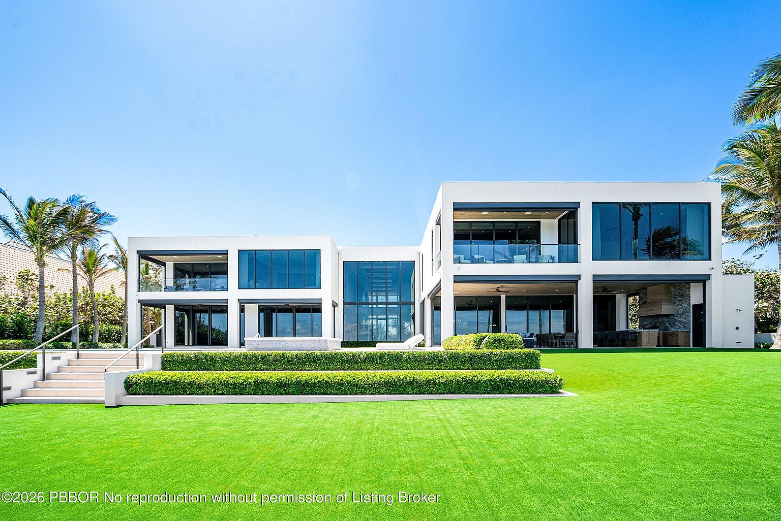 This striking modern residence features a clean, geometric architectural design with expansive glass walls and a bright white facade. The rear of the home opens onto a meticulously manicured lawn, tiered landscaping with structured hedges, and a set of wide concrete steps leading to the main living level. The composition captures the home's seamless indoor-outdoor transition, highlighting its contemporary aesthetic and generous outdoor living space under a clear blue sky.