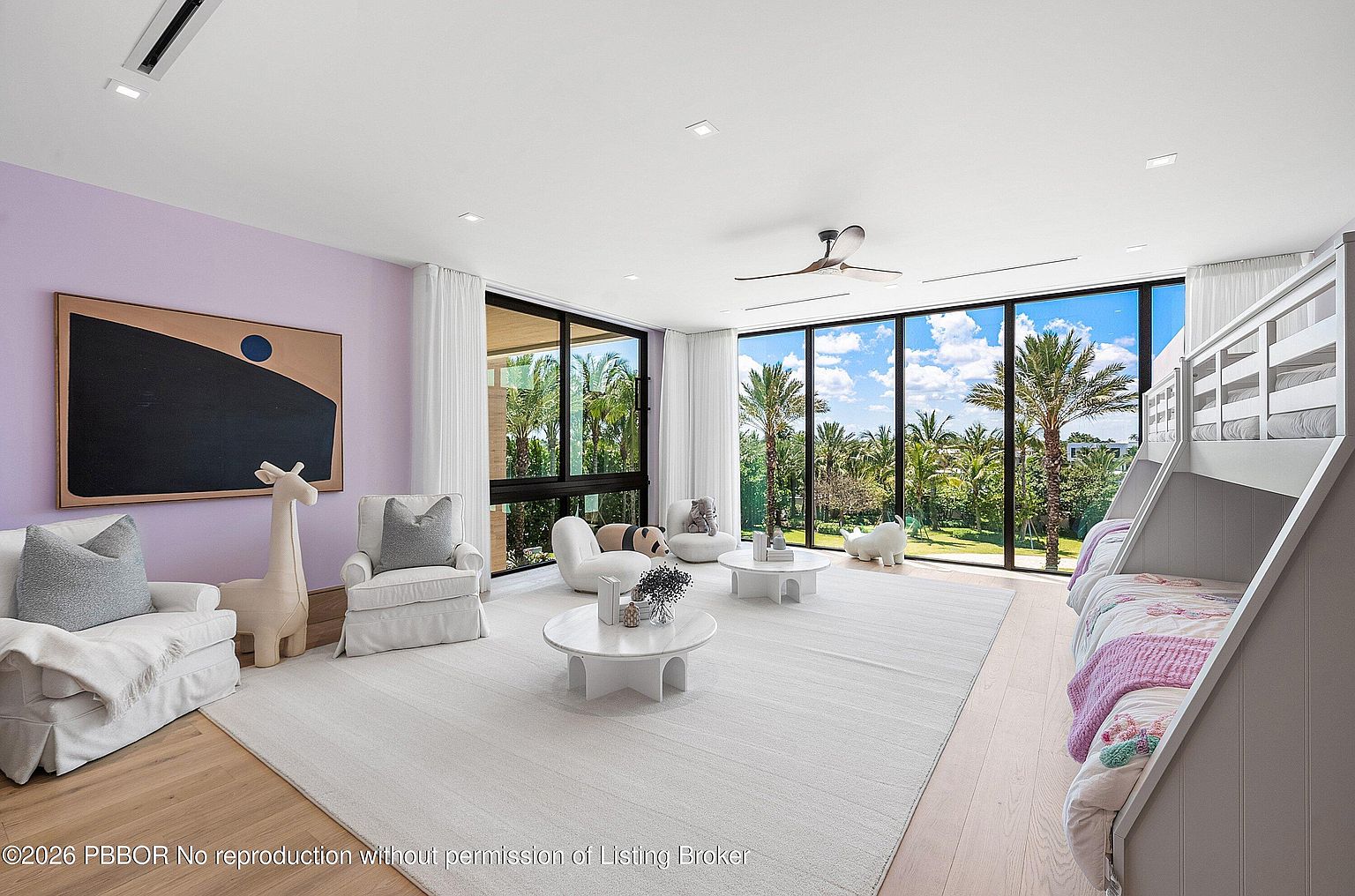 This spacious, light-filled bedroom features a soft lavender accent wall and a large bunk bed setup, making it an ideal children's room. The space is anchored by a large, light-colored area rug and includes two circular coffee tables and comfortable lounge seating. Expansive floor-to-ceiling windows provide an unobstructed view of the lush, tropical greenery outside, creating a bright and airy atmosphere.