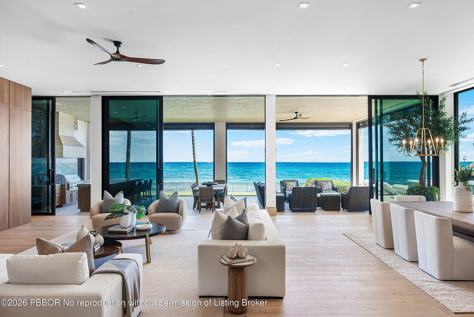 This expansive, open-concept living area features a seamless transition to a covered outdoor patio through large glass sliding doors that frame a stunning ocean view. The interior is decorated in a sophisticated, neutral palette with light wood flooring, plush cream-colored seating, and a modern chandelier over the dining area. The space feels airy and luxurious, emphasizing a strong connection between the refined indoor living space and the coastal environment.