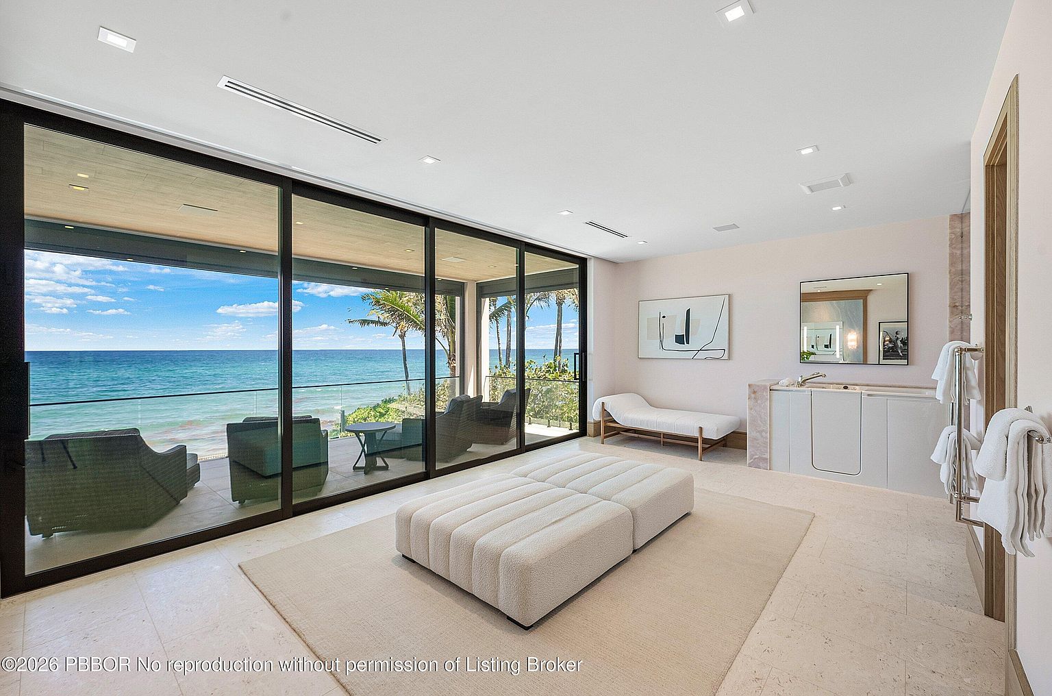 This luxurious primary bathroom features a minimalist aesthetic with light-toned stone flooring and a large, plush ottoman centered on a neutral area rug. A wall of floor-to-ceiling glass sliding doors provides an expansive, unobstructed view of the ocean and a private balcony. The space includes a sleek vanity area with a built-in tub, a chaise lounge for relaxation, and modern recessed lighting, creating a serene and high-end spa-like atmosphere.