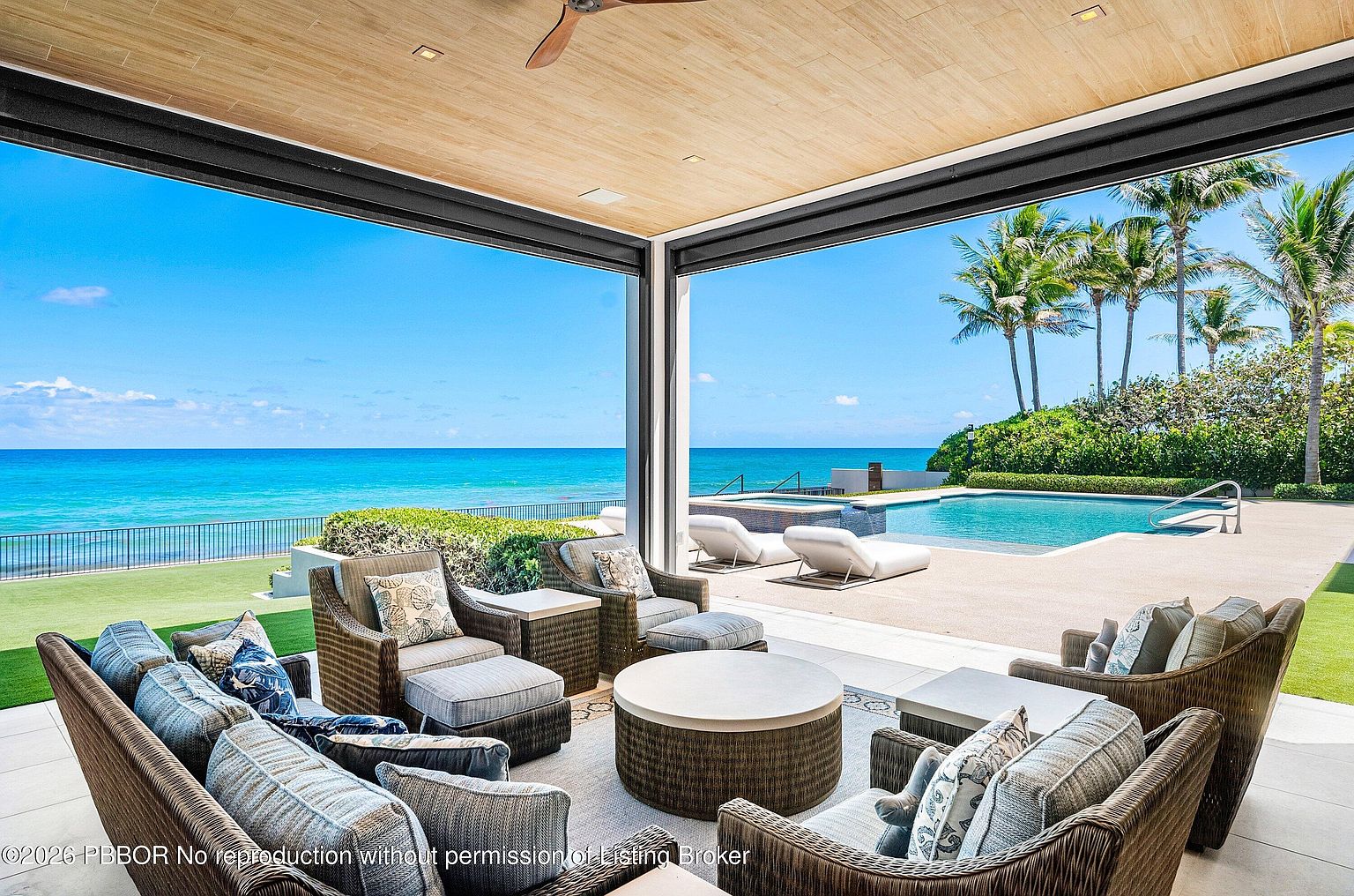 This luxurious covered patio offers a seamless transition to an outdoor living space, featuring a comfortable seating area with wicker furniture and plush cushions arranged around a central circular coffee table. The open-air design provides an unobstructed, panoramic view of the ocean, a pristine swimming pool, and a manicured lawn. The space is defined by a light wood-paneled ceiling and a clean, modern aesthetic that emphasizes indoor-outdoor living.