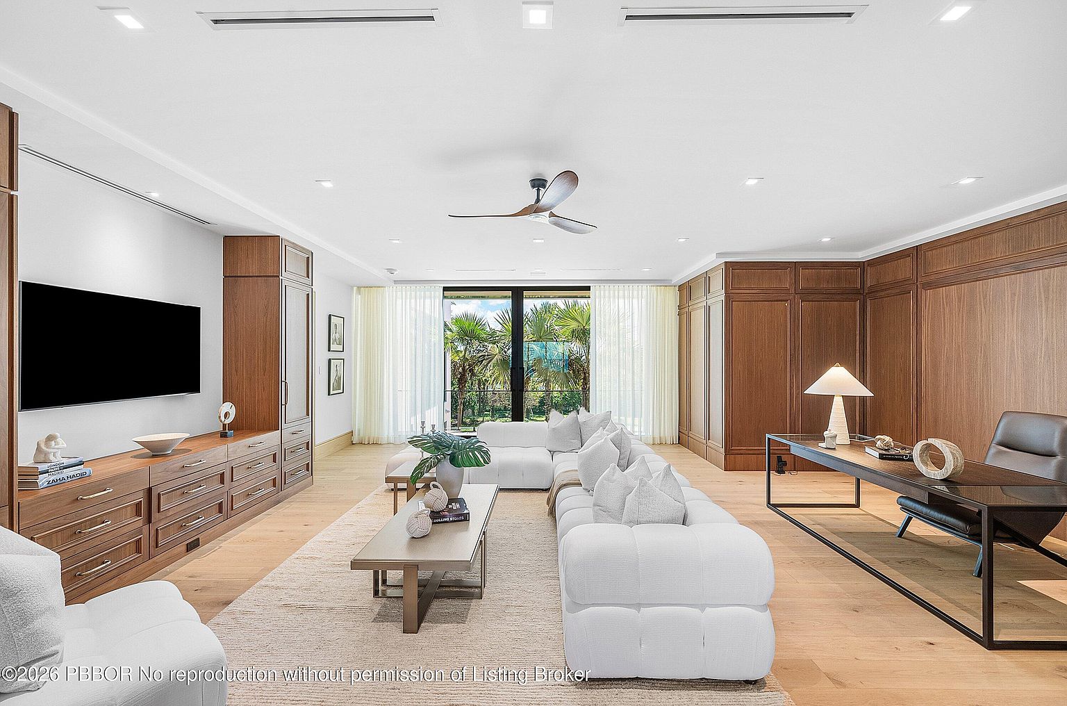 This expansive, modern living room features a large white sectional sofa arranged around a central coffee table, set against a backdrop of warm wood-paneled walls and built-in cabinetry. A sleek desk area is integrated into the space, while floor-to-ceiling glass doors at the far end provide a clear view of lush greenery outside. The room is illuminated by recessed lighting and a ceiling fan, creating a sophisticated and airy atmosphere.