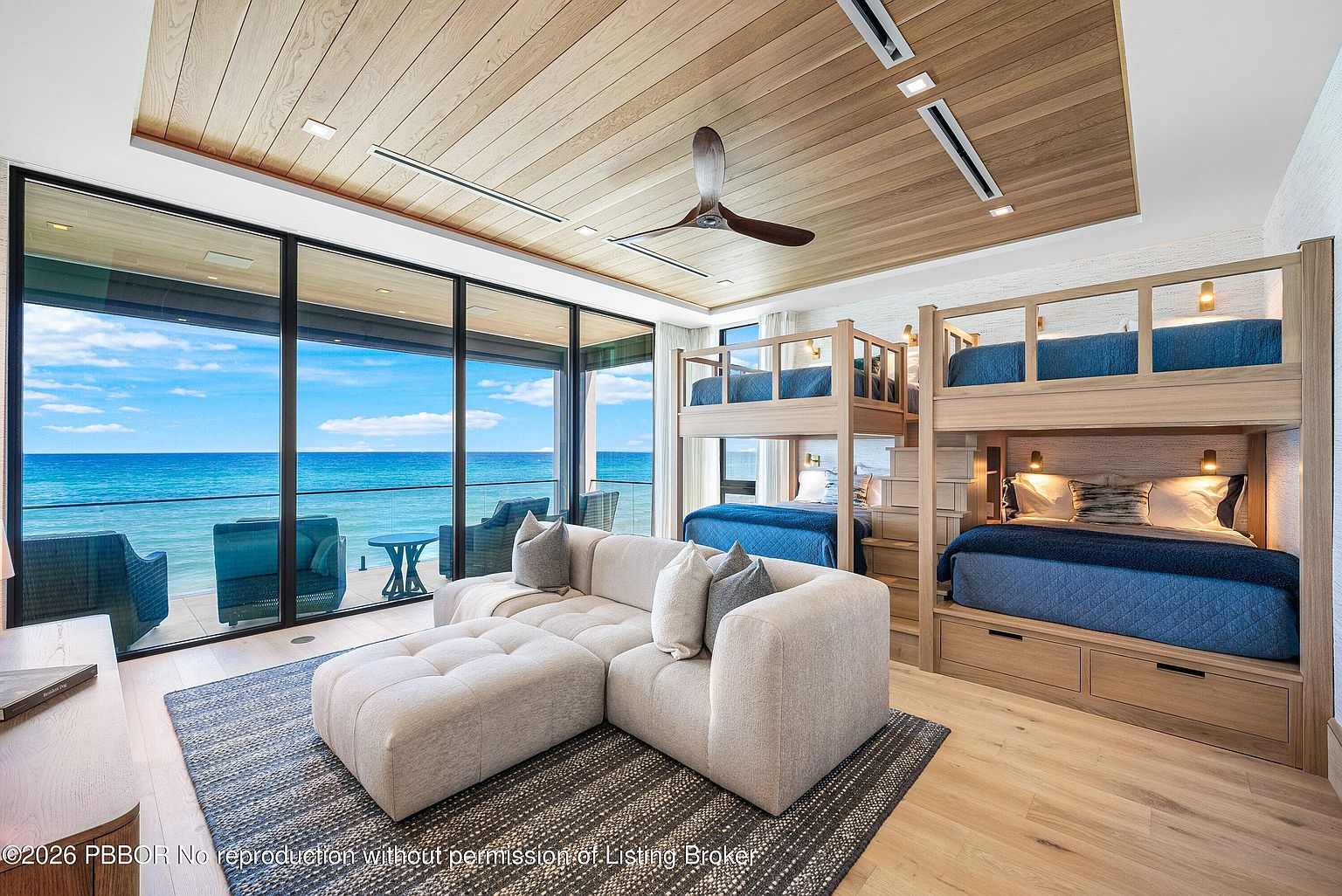 This luxurious bedroom features a unique bunk bed configuration with integrated stairs and built-in storage, finished in light wood. A large, plush sectional sofa sits on a textured area rug in the center of the room, facing floor-to-ceiling windows that provide an expansive, unobstructed view of the ocean. The space is accented by a wood-paneled ceiling with recessed lighting and a modern ceiling fan, creating a serene and high-end coastal retreat.