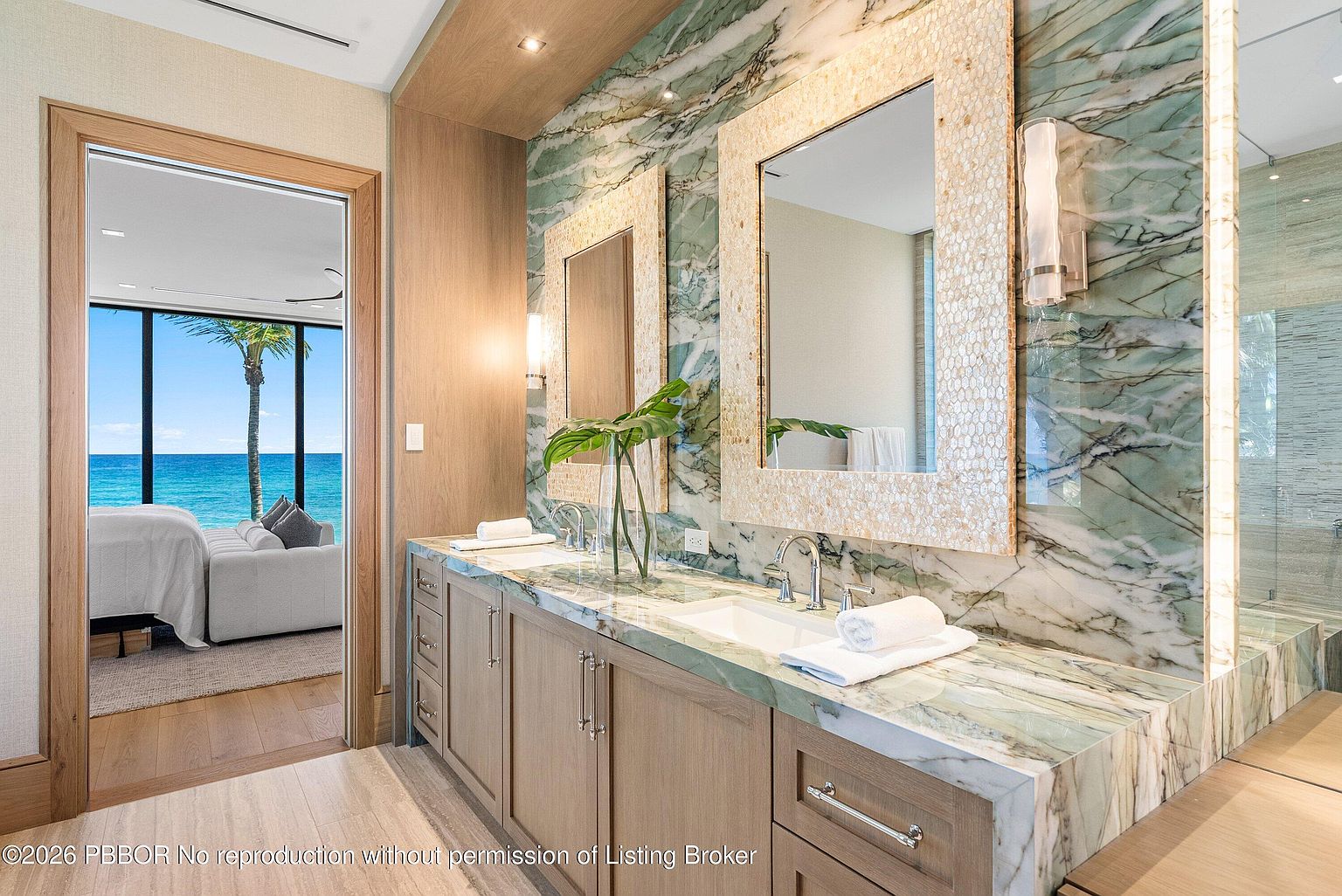 This luxurious primary bathroom features a dual-vanity with light wood cabinetry and a striking green-veined stone countertop that extends up the wall as a backsplash. Two rectangular mirrors with textured, light-colored frames are mounted above the sinks, complemented by modern wall sconces. A doorway provides a direct, scenic view into an adjacent bedroom with floor-to-ceiling windows overlooking the ocean.