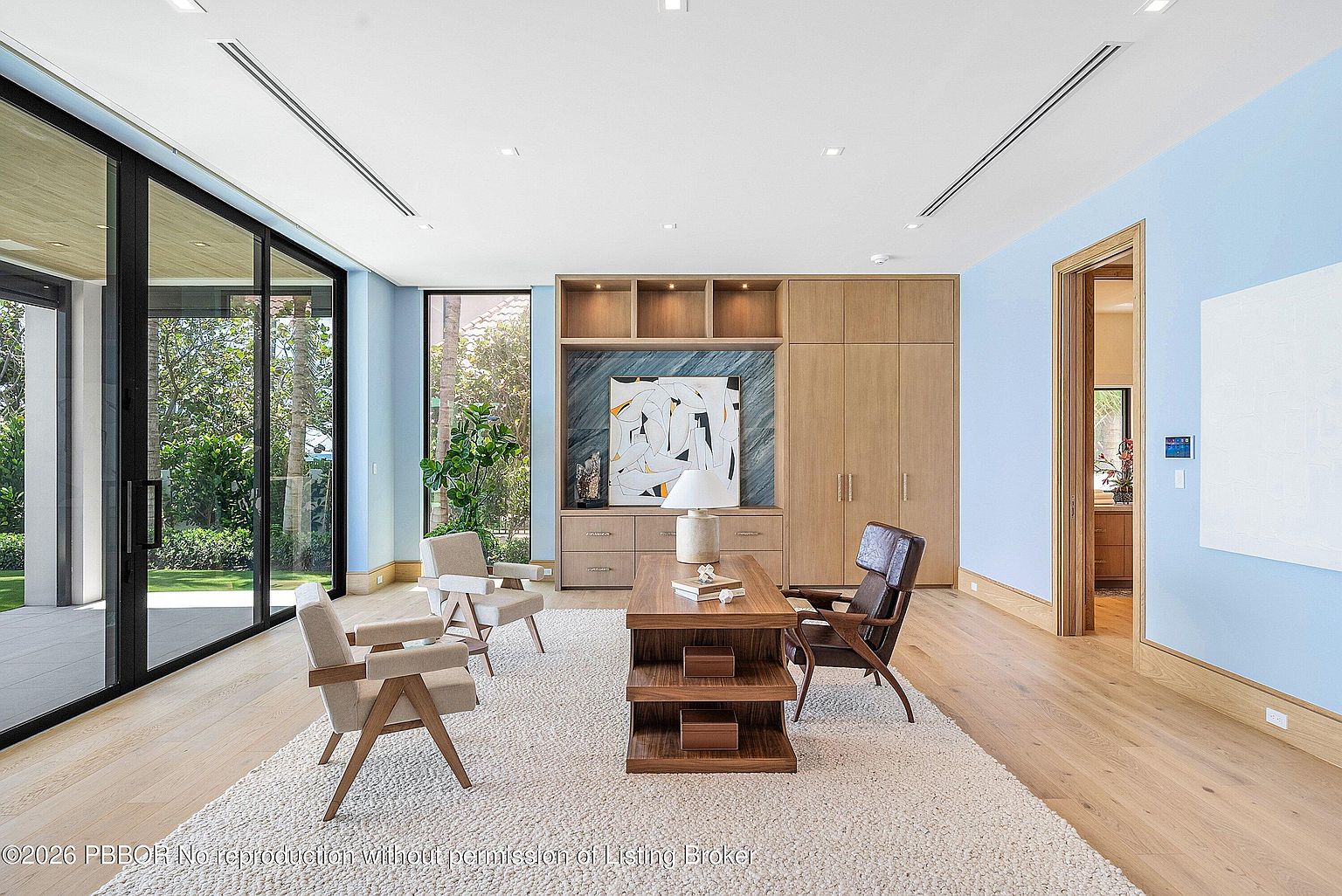 This sophisticated home office features light blue walls, warm wood flooring, and a large desk positioned centrally on a textured area rug. A custom built-in unit with integrated shelving and cabinetry provides ample storage, while floor-to-ceiling glass doors offer a seamless view of the lush outdoor greenery. The room is bathed in natural light, creating a serene and professional atmosphere suitable for focused work.