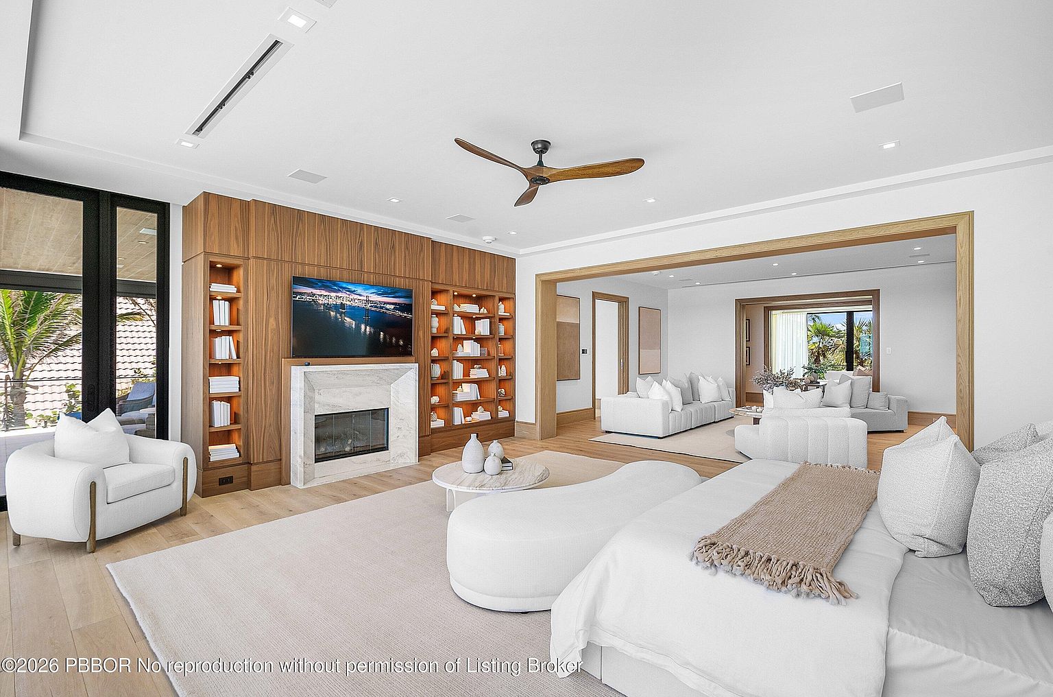 This expansive primary bedroom features a sophisticated design with light wood flooring and a large, integrated wood-paneled wall unit that incorporates a television and a stone-clad fireplace. The room is furnished with a plush white bed, a curved ottoman, and a comfortable armchair, all arranged on a neutral area rug. A wide opening leads into an adjacent sitting area, creating a seamless flow between spaces while maintaining an airy, open-concept feel.