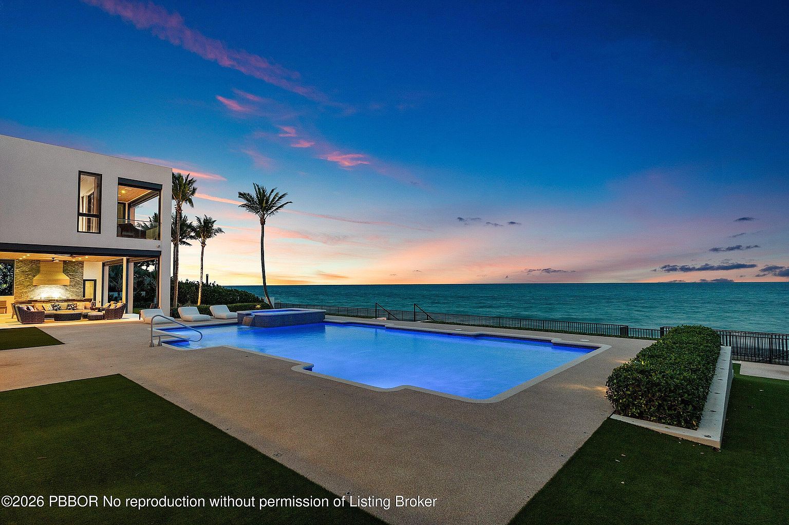 This stunning waterfront property features a modern, multi-level home with a large, rectangular swimming pool and integrated spa overlooking the ocean. The expansive patio area includes a covered outdoor living space with a stone-clad wall, while the horizon is captured during a vibrant sunset. The scene conveys a luxurious, tranquil atmosphere perfect for high-end coastal living.