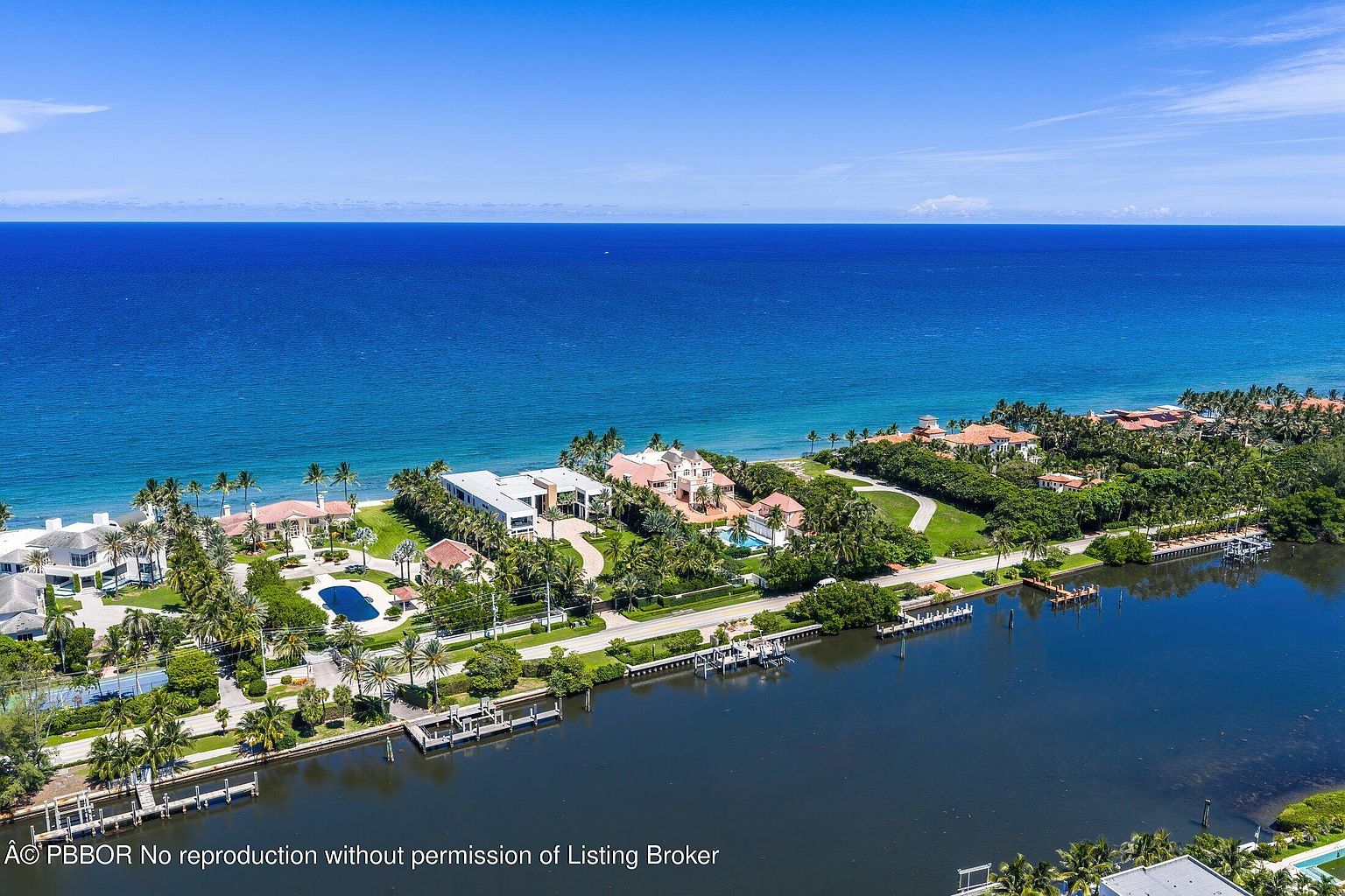 This high-altitude aerial view captures a luxurious coastal residential enclave situated between a calm inland waterway and the vast Atlantic Ocean. The scene showcases several expansive estates with manicured lawns, private swimming pools, and lush tropical landscaping, all connected by a winding road. The perspective emphasizes the prime waterfront location, highlighting the seamless transition from the deep blue sea to the private docks along the canal.