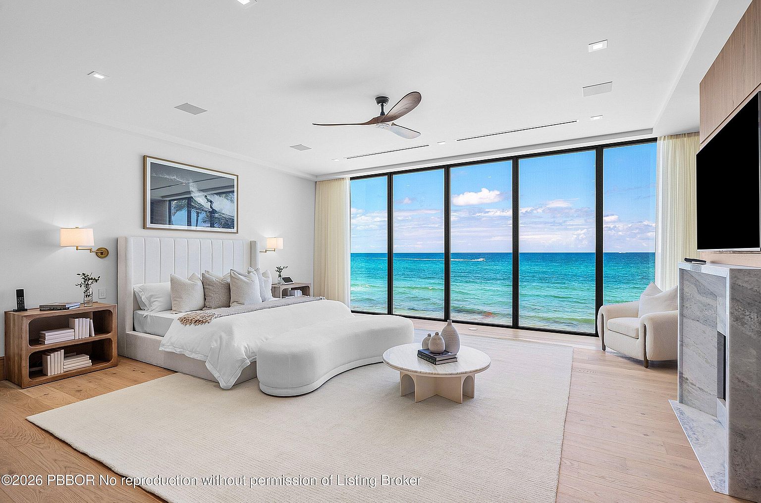 This luxurious primary bedroom features a neutral color palette and a minimalist aesthetic, centered around a plush bed with a tall, upholstered headboard. A wall of floor-to-ceiling windows provides an expansive, unobstructed view of the turquoise ocean, filling the room with natural light. The space is furnished with a curved bench, a circular stone coffee table, and a comfortable armchair positioned near a stone-clad fireplace surround.