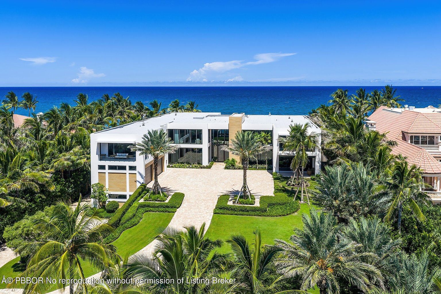 This high-angle aerial view captures a modern, multi-level luxury residence situated directly along the coastline. The home features a clean, white geometric facade with extensive glass elements, a paved motor court, and lush tropical landscaping. The expansive blue ocean serves as a dramatic backdrop, emphasizing the property's prime waterfront location.