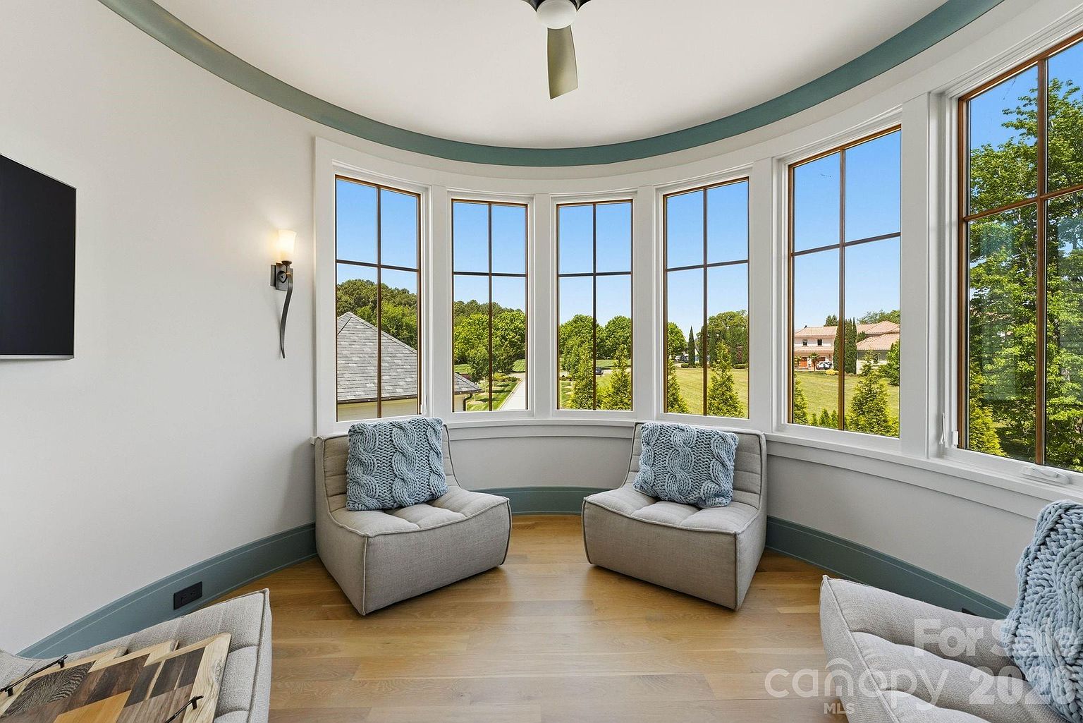 This bright, curved seating area features a series of large windows that provide expansive views of the surrounding landscape. The space is furnished with two comfortable, low-profile armchairs accented with textured blue pillows, set against a backdrop of light wood flooring and soft-toned walls. A ceiling fan and a wall-mounted light fixture add functional lighting to this serene, sun-drenched nook.