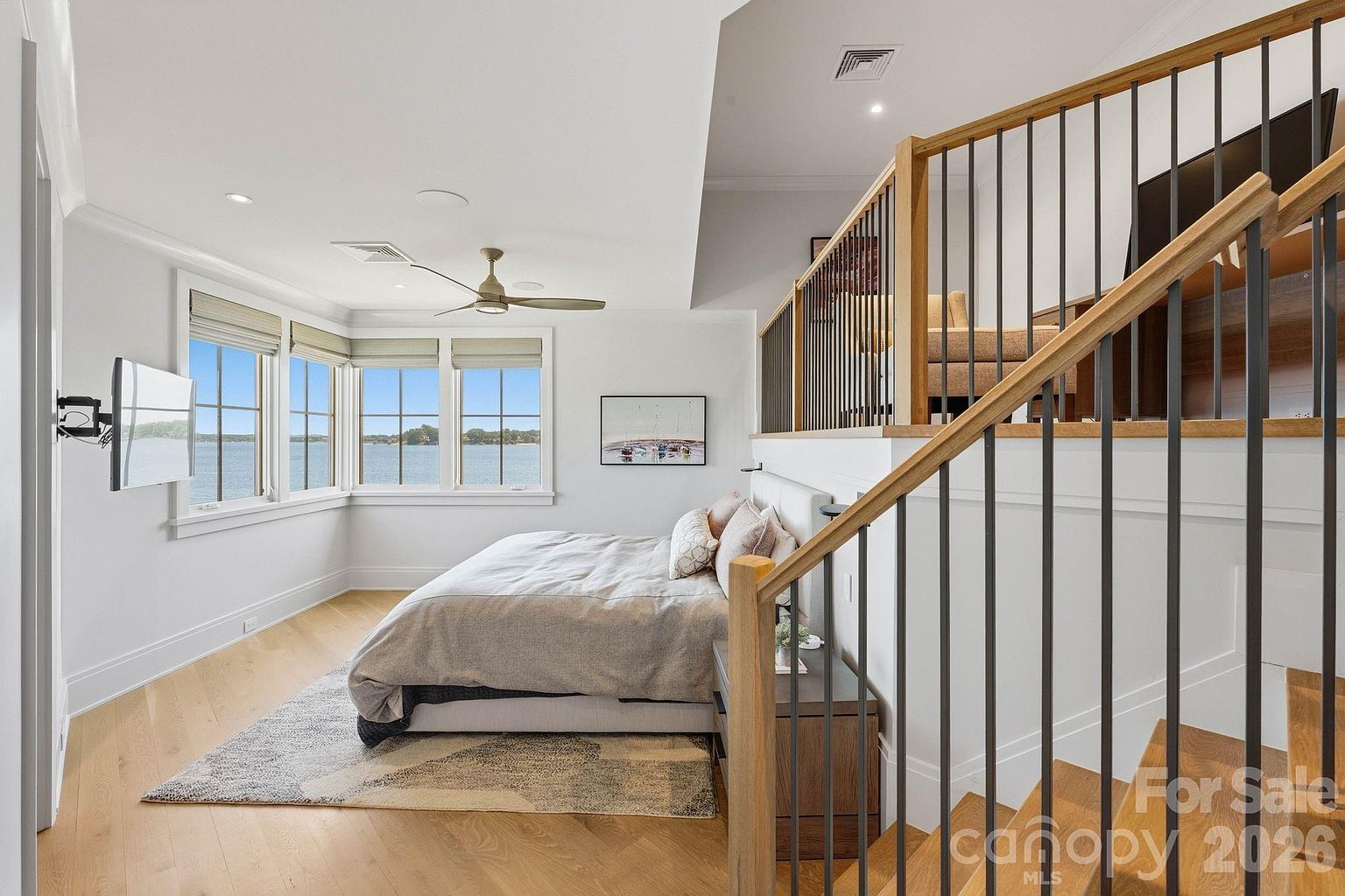This primary bedroom features a sophisticated, multi-level layout with a lofted area overlooking the sleeping space. The room is anchored by a large bed positioned against a low wall, with expansive windows providing a panoramic view of the water. Light wood flooring, neutral tones, and a modern ceiling fan create a serene and airy atmosphere, while a staircase with dark metal railings adds architectural interest.