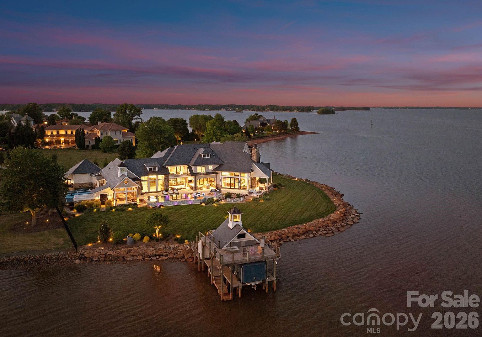 This stunning aerial view captures a luxurious waterfront estate at dusk, showcasing its expansive architecture and prime location on a point of land. The property features a multi-level residence with illuminated interiors, a sprawling lawn, a private swimming pool, and a charming boat dock extending into the calm water. The dramatic sunset sky provides a picturesque backdrop, emphasizing the home's secluded and high-end appeal.