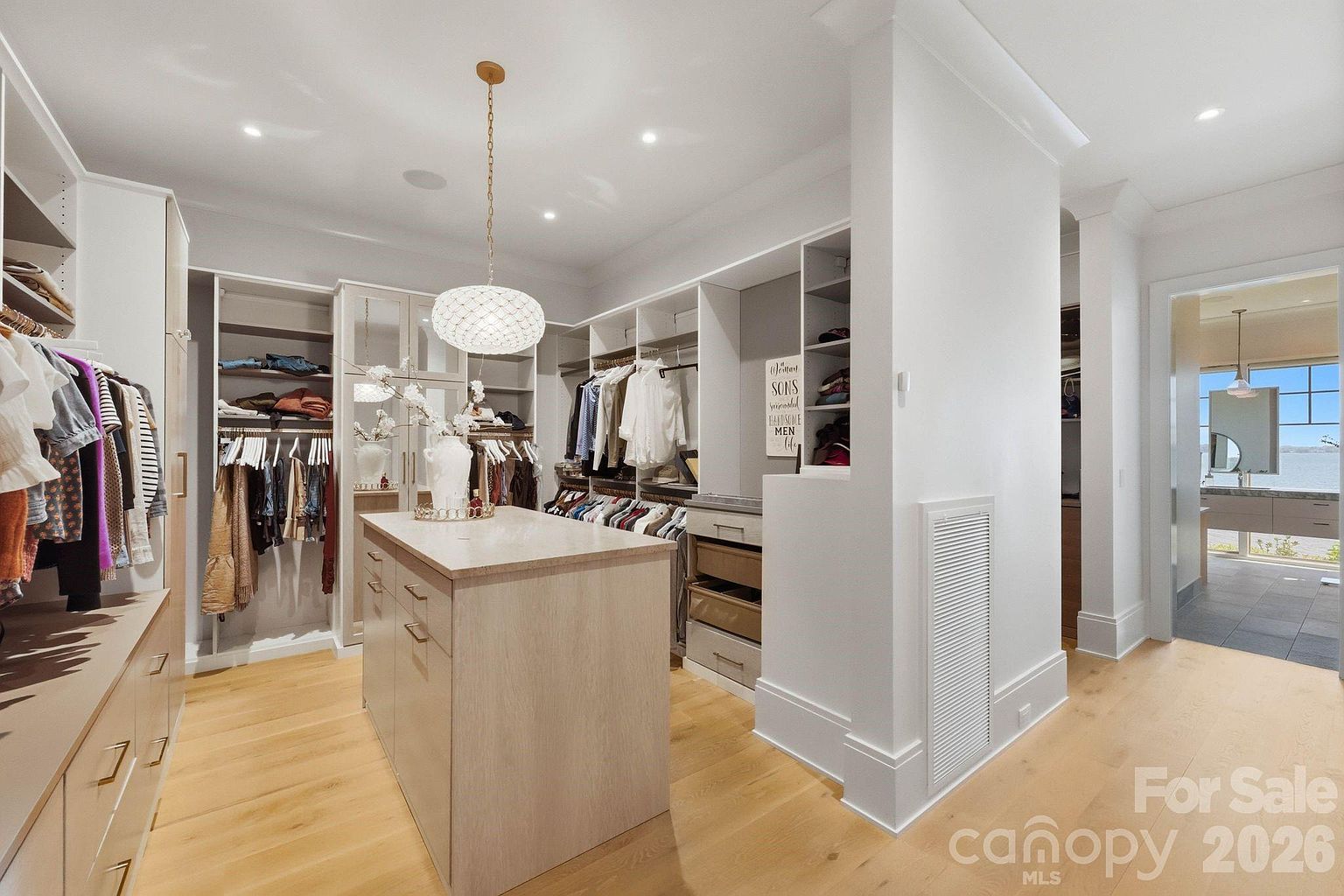 This spacious, well-organized walk-in closet features light wood cabinetry, a central island with drawers, and ample hanging space for clothing. A decorative pendant light hangs above the island, while the neutral color palette and light wood flooring create a bright, airy atmosphere. The layout includes a transition area leading toward a bathroom with a view of the water, emphasizing a luxurious and functional design.
