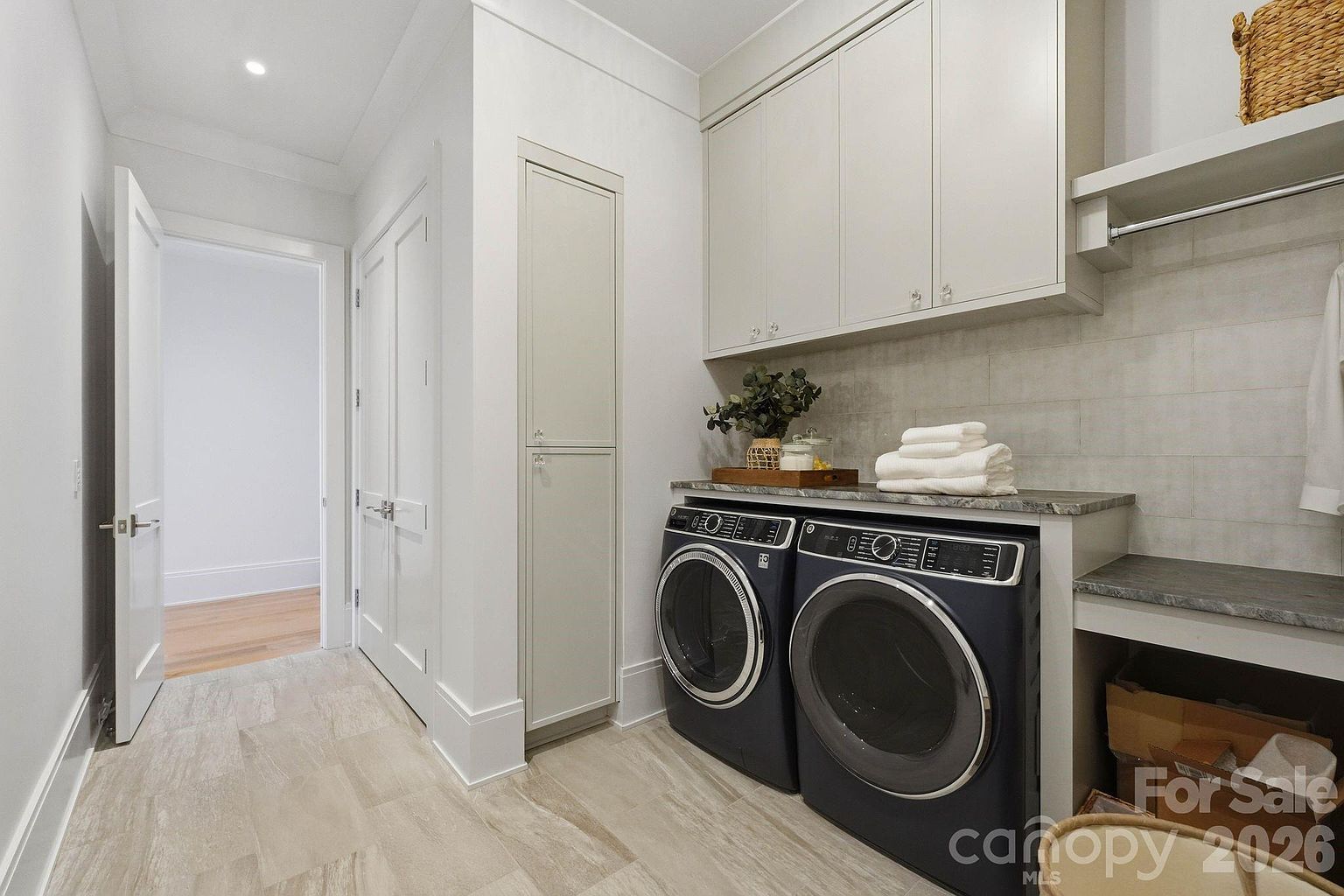 This clean and modern laundry room features dark-toned front-loading washer and dryer units set beneath a stone countertop. The space is equipped with ample upper cabinetry for storage, a tall utility cabinet, and a hanging rod for garments. The neutral color palette and light-toned flooring create a bright, organized, and functional utility area.